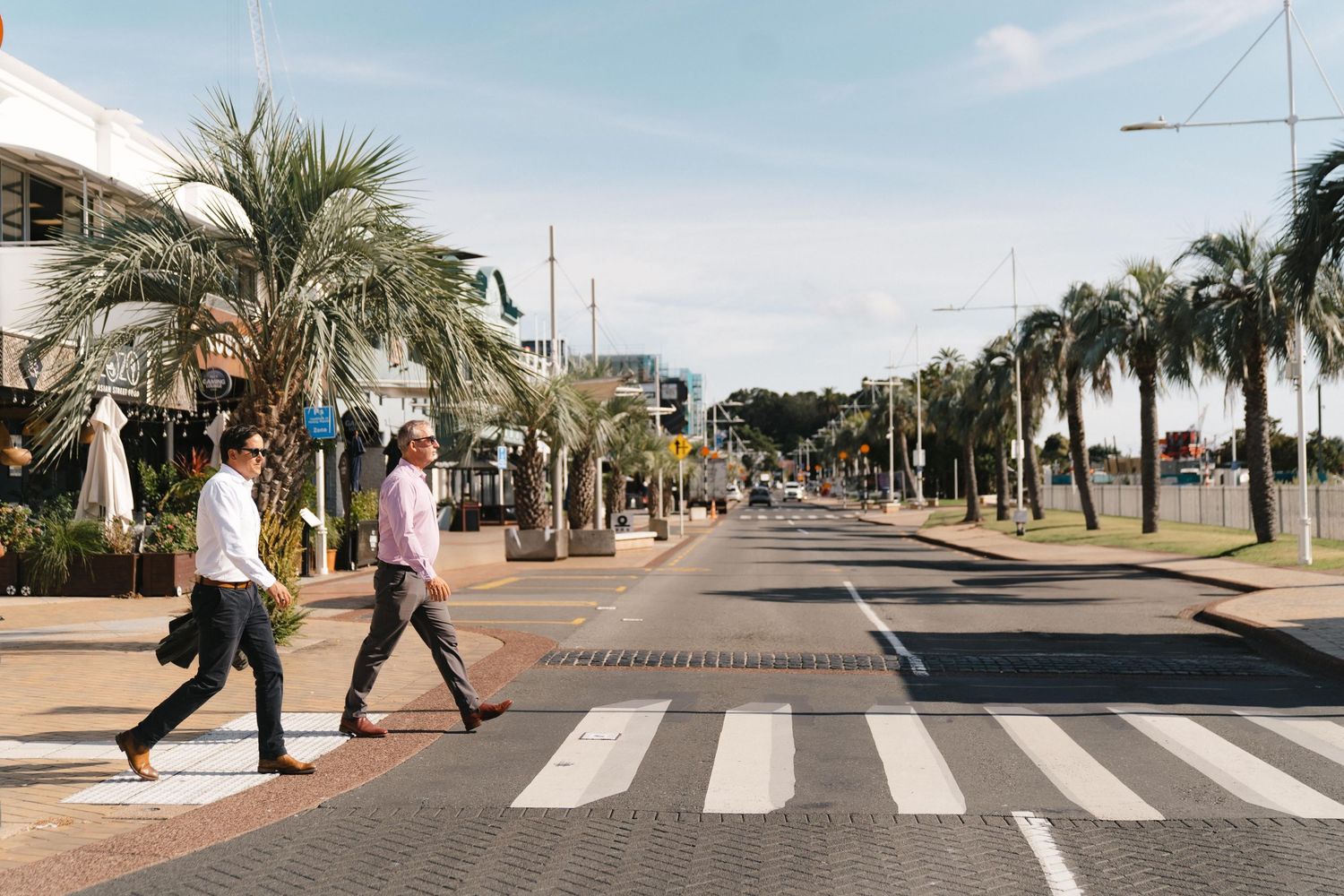 People walking across a zebra crossing on a palm-lined street in a tropical location.