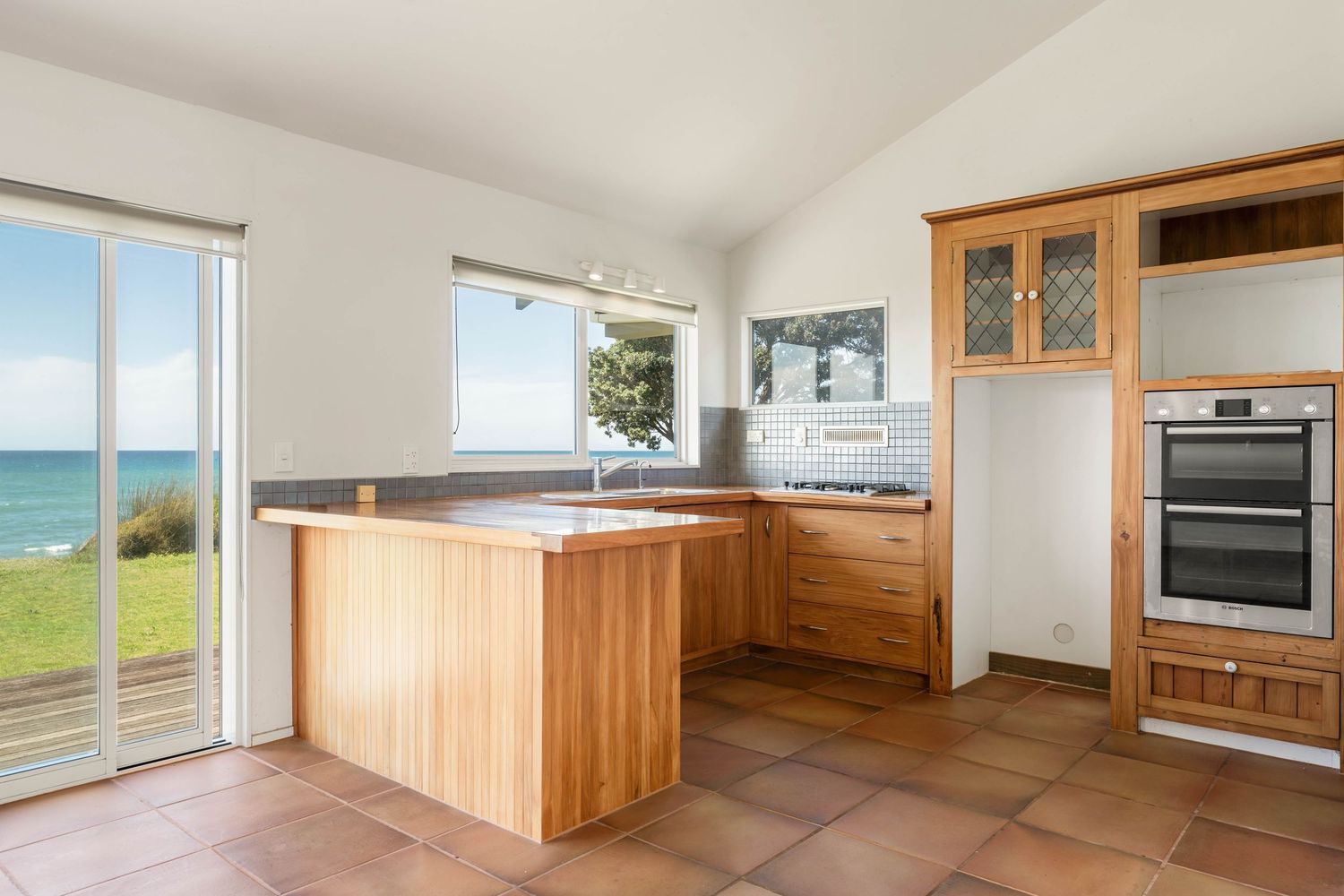 Modern kitchen with wooden cabinets and ocean view through large glass doors featuring terracotta tile flooring.