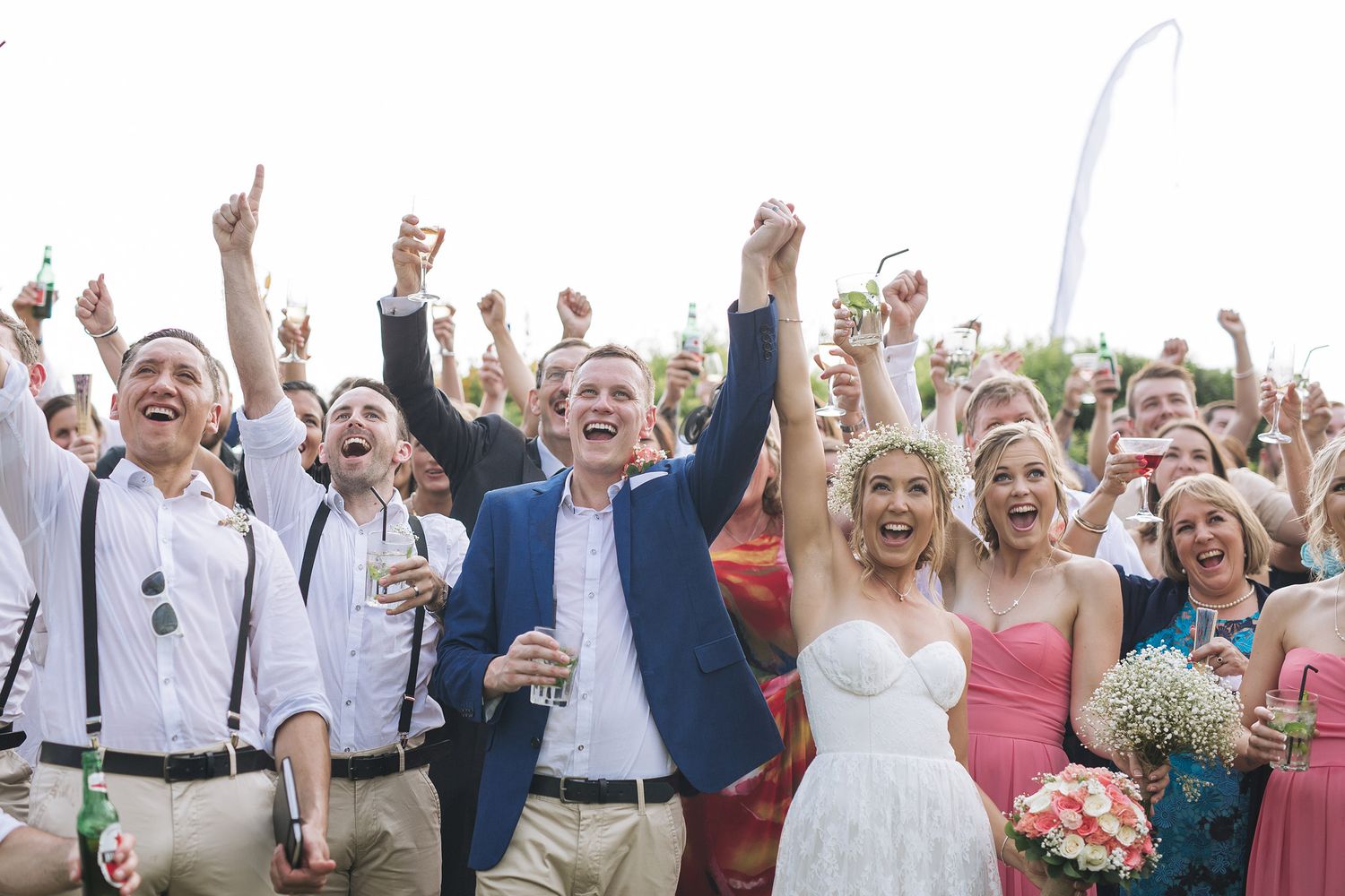 Wedding party and guests celebrating with raised arms and joyful expressions outdoors.