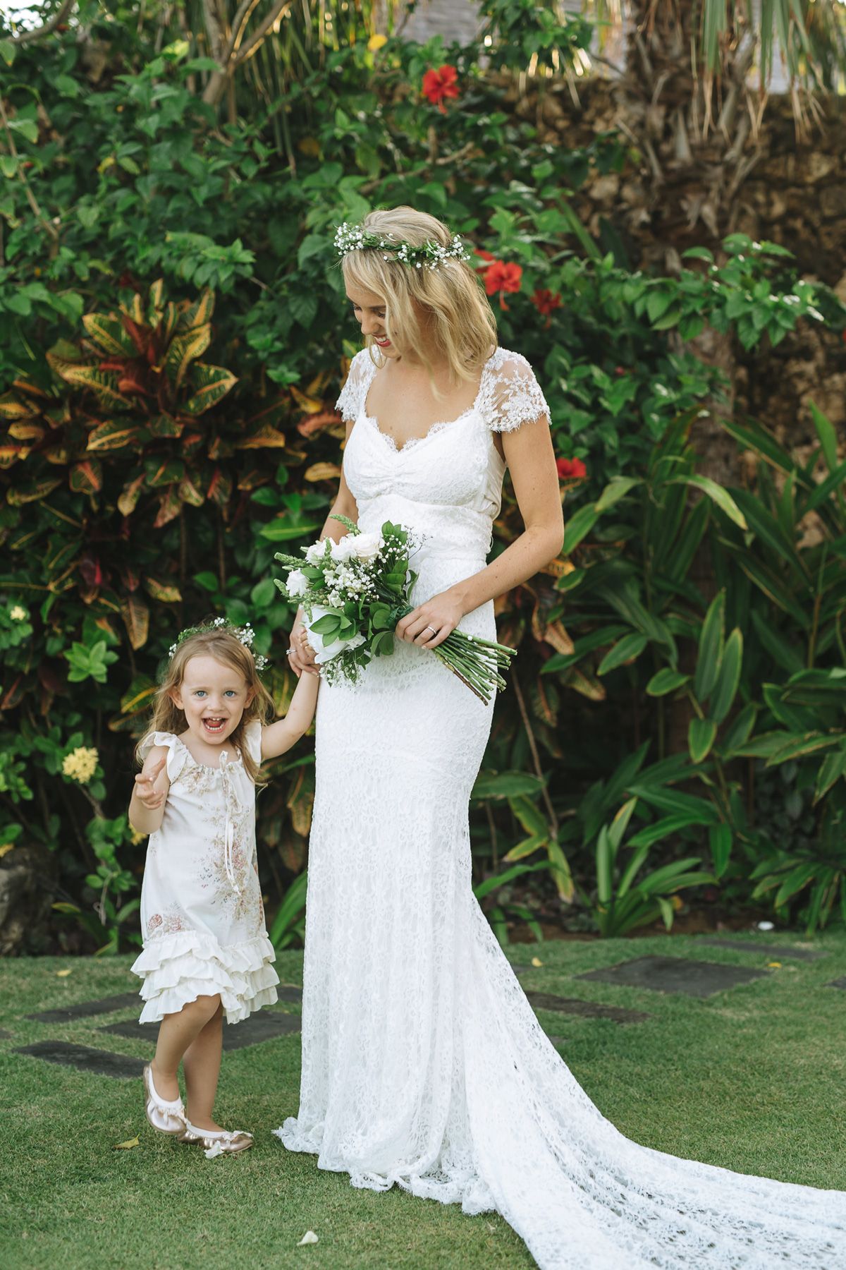 A young girl in white dress shares a joyful moment with someone in a lace wedding gown in a tropical garden.