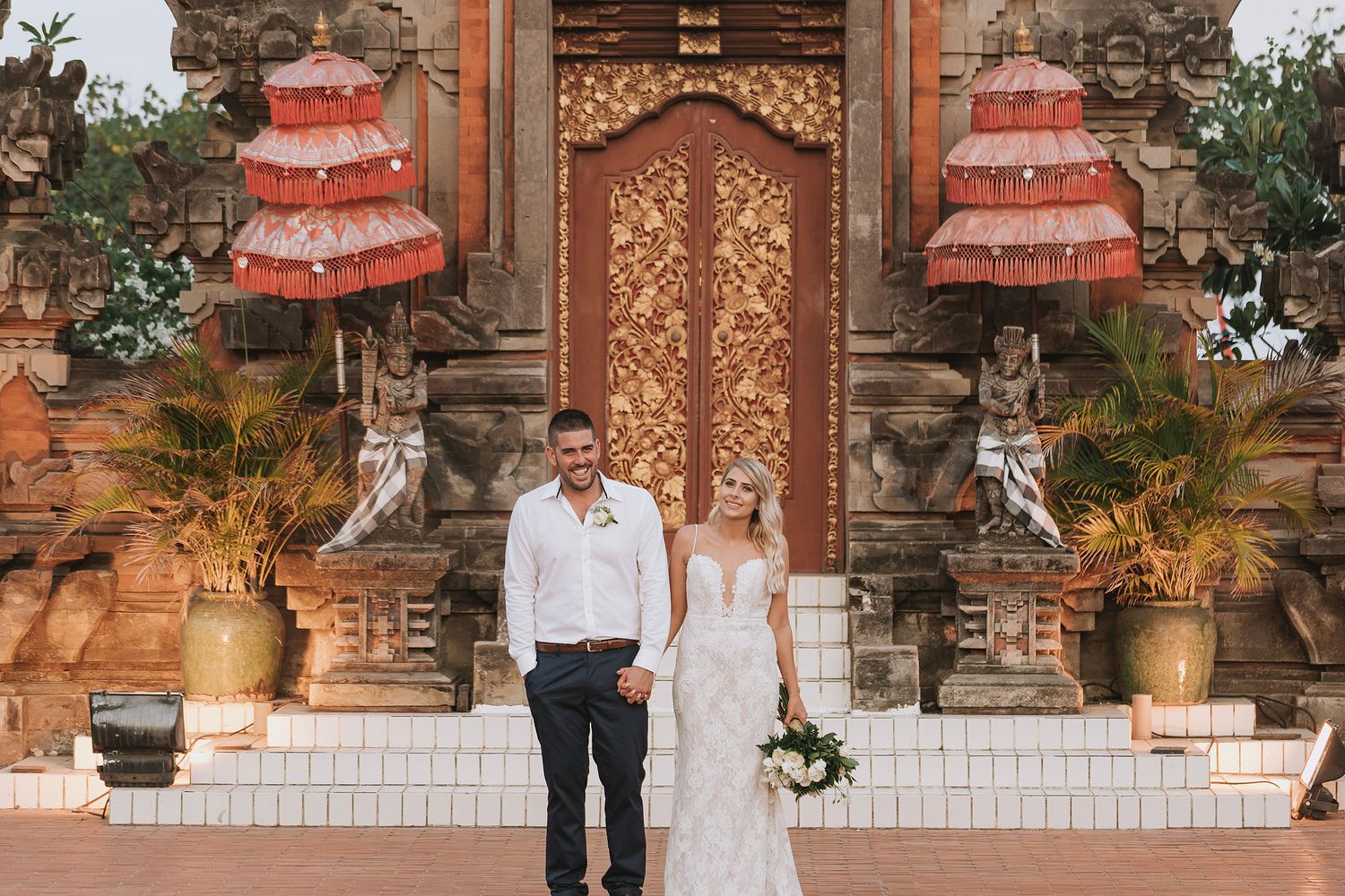 Wedding ceremony scene in front of ornate Balinese temple gates with traditional red tiered roofs.