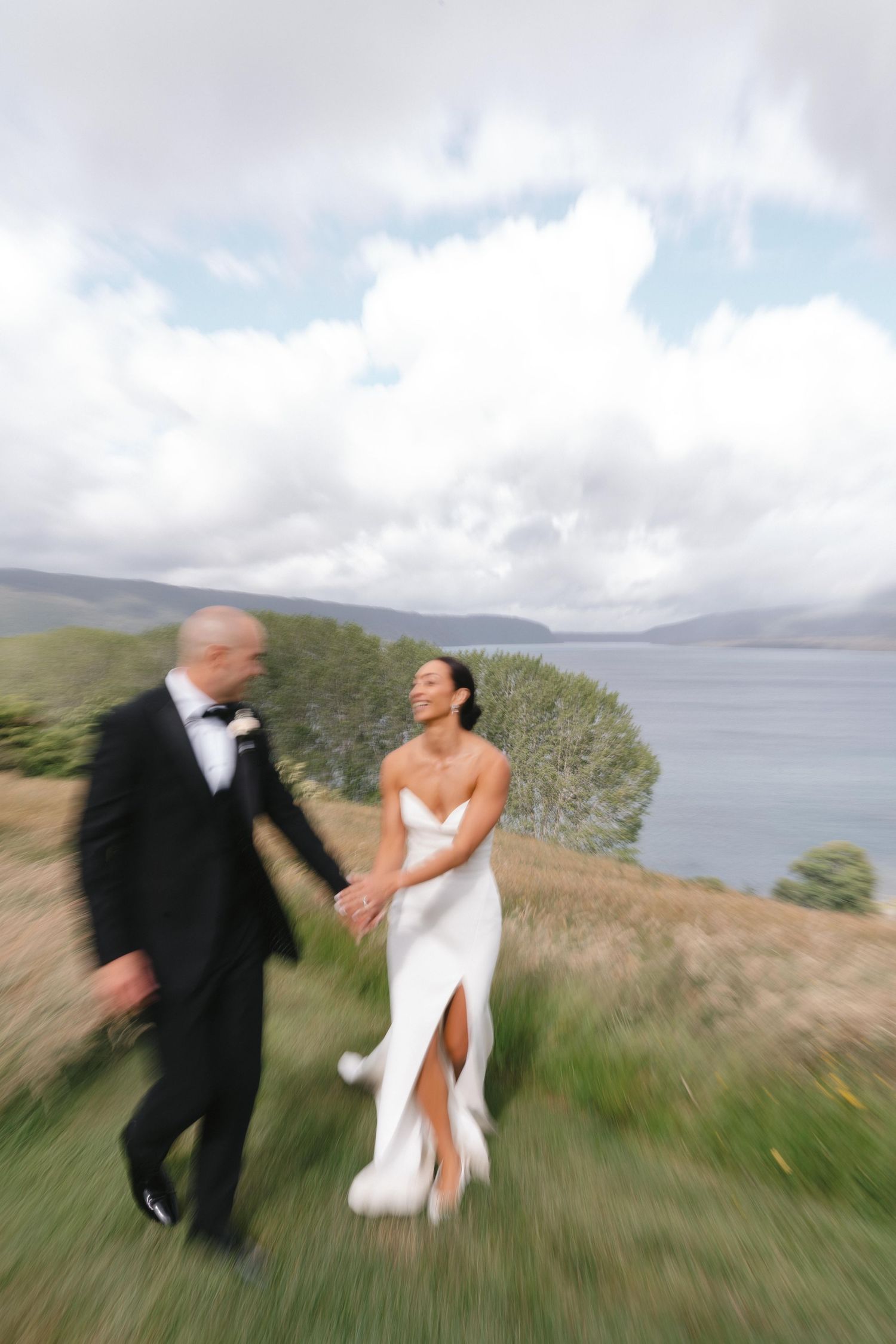 Couple running playfully through tall grass on a coastal cliff with water views.