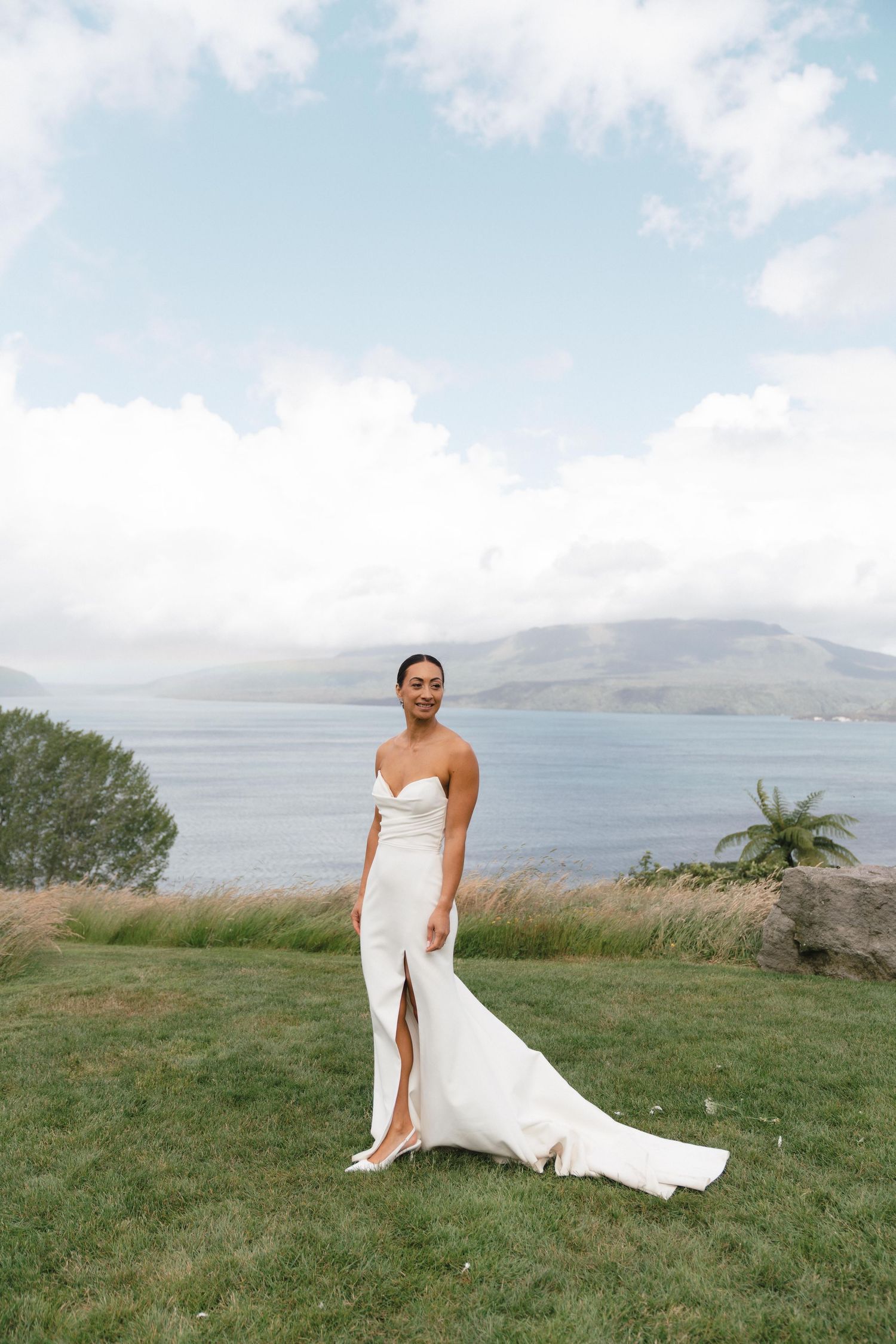 Bride in white dress with high slit posing on a grassy cliff overlooking the ocean.