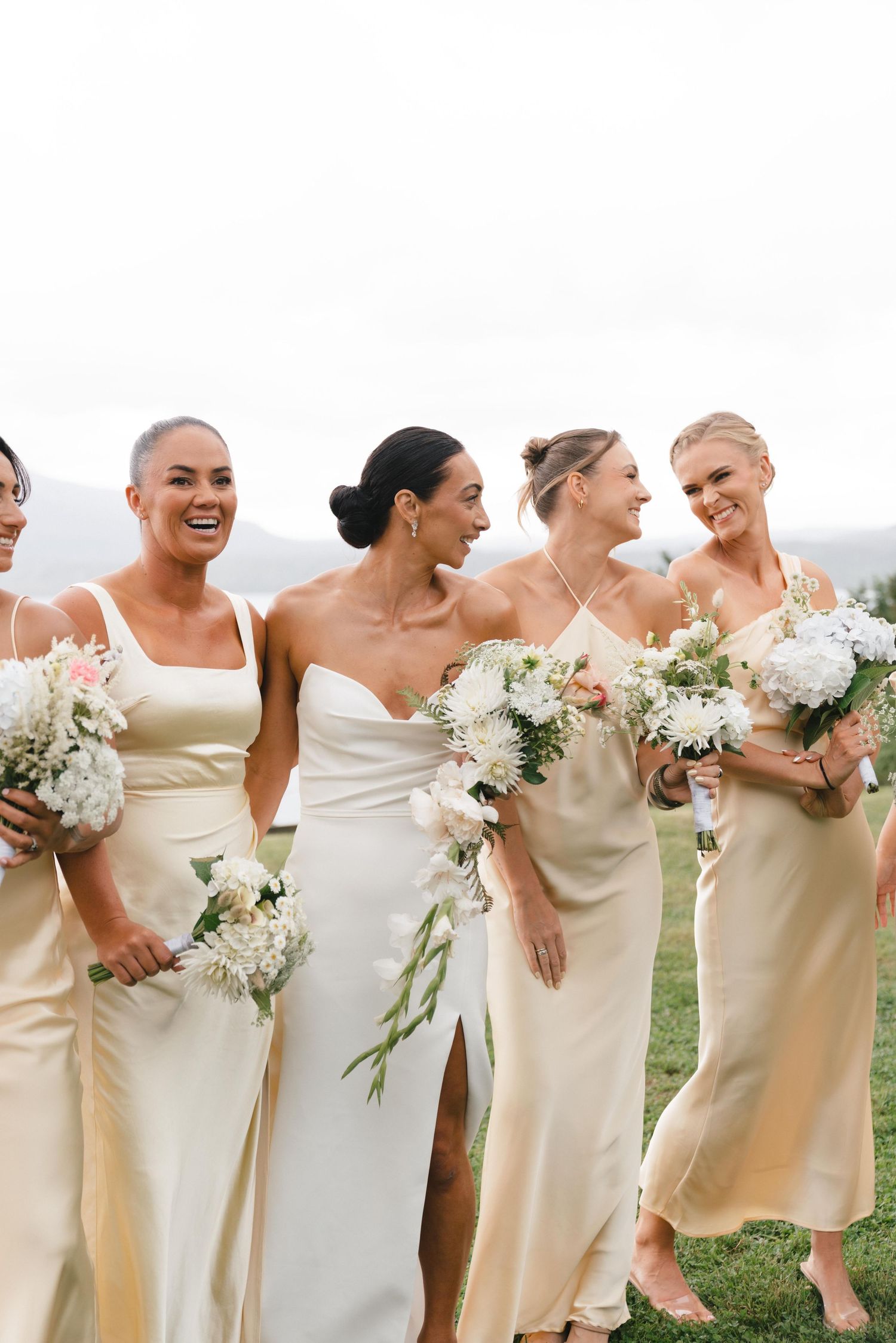 Bridal party in champagne silk dresses share laughter while holding white bouquets.