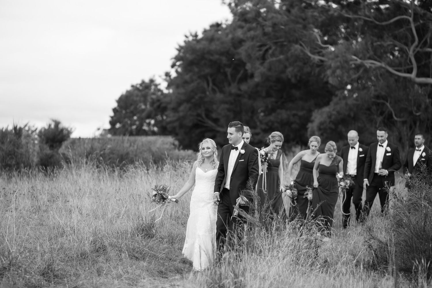 A black and white wedding photo shows a bridal party standing together in a rustic field with tall grass and trees.