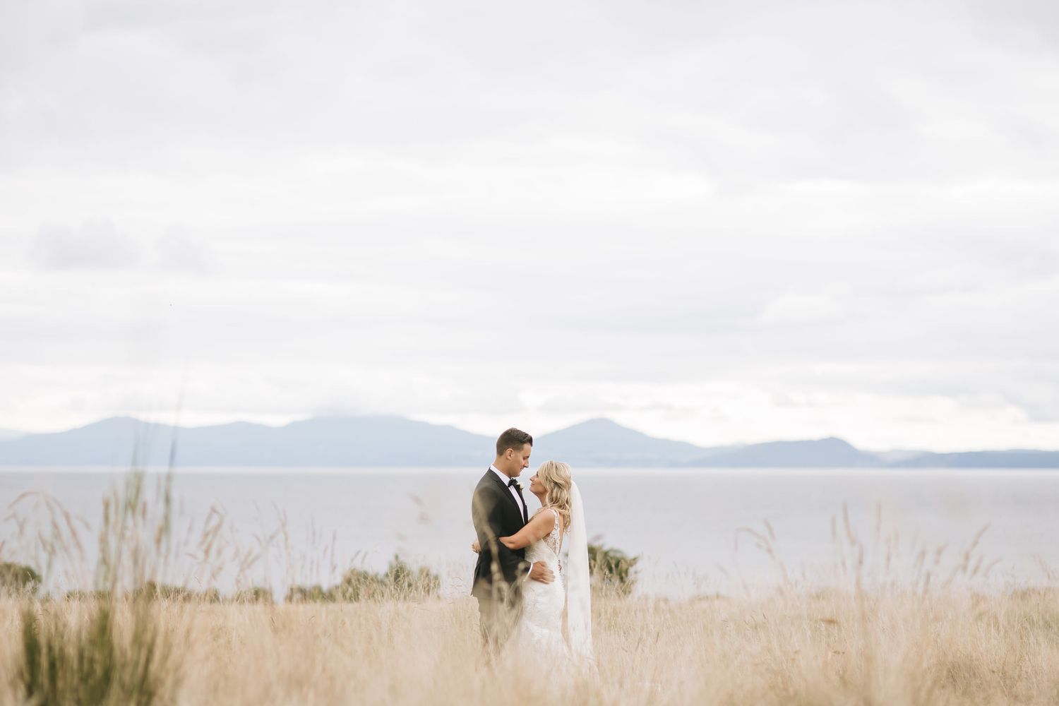 A loving couple shares a tender moment in a grassy field overlooking a scenic mountain lake view.