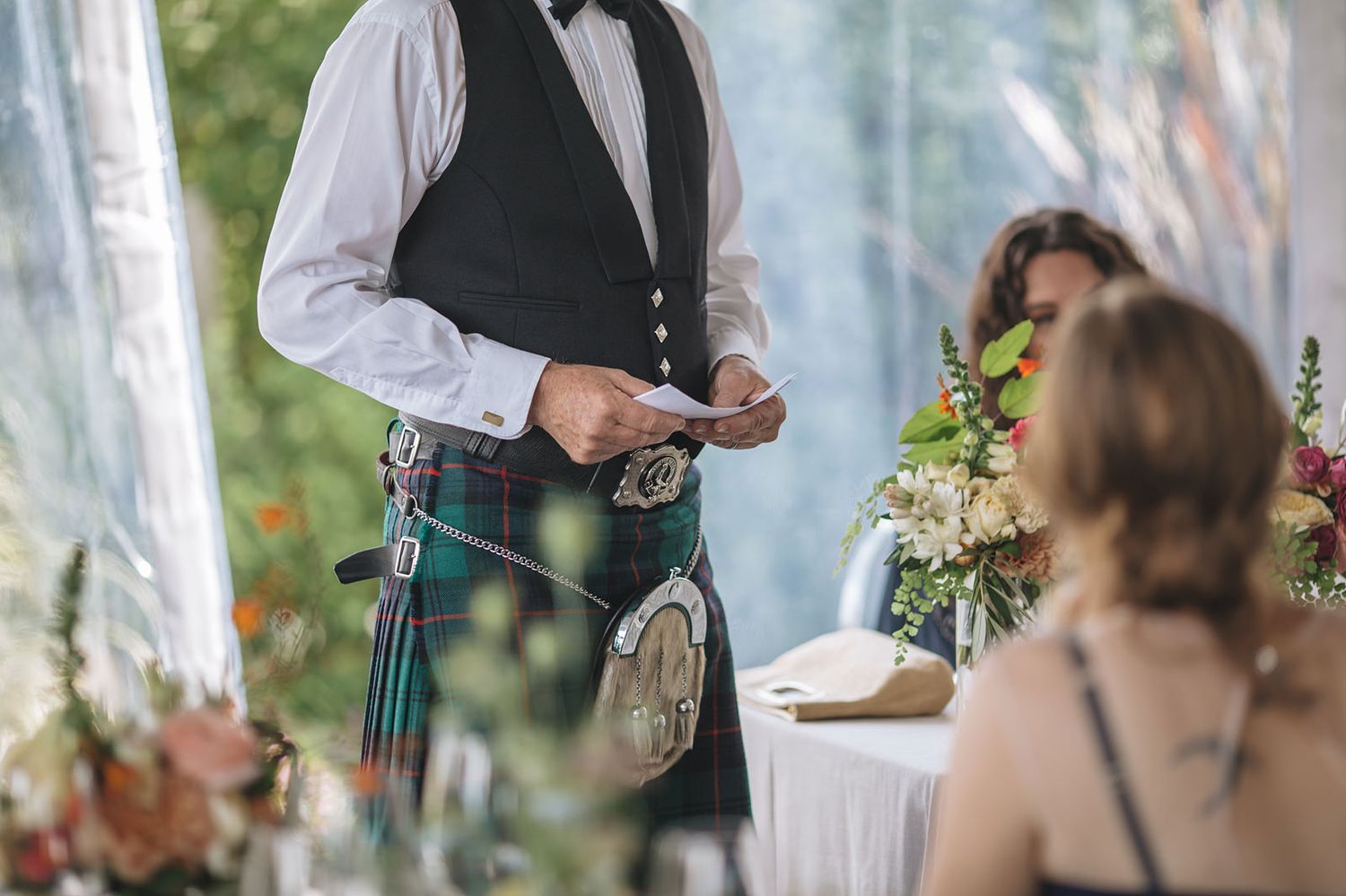 Scottish wedding server in traditional kilt and vest takes drink orders inside an elegant outdoor reception tent.