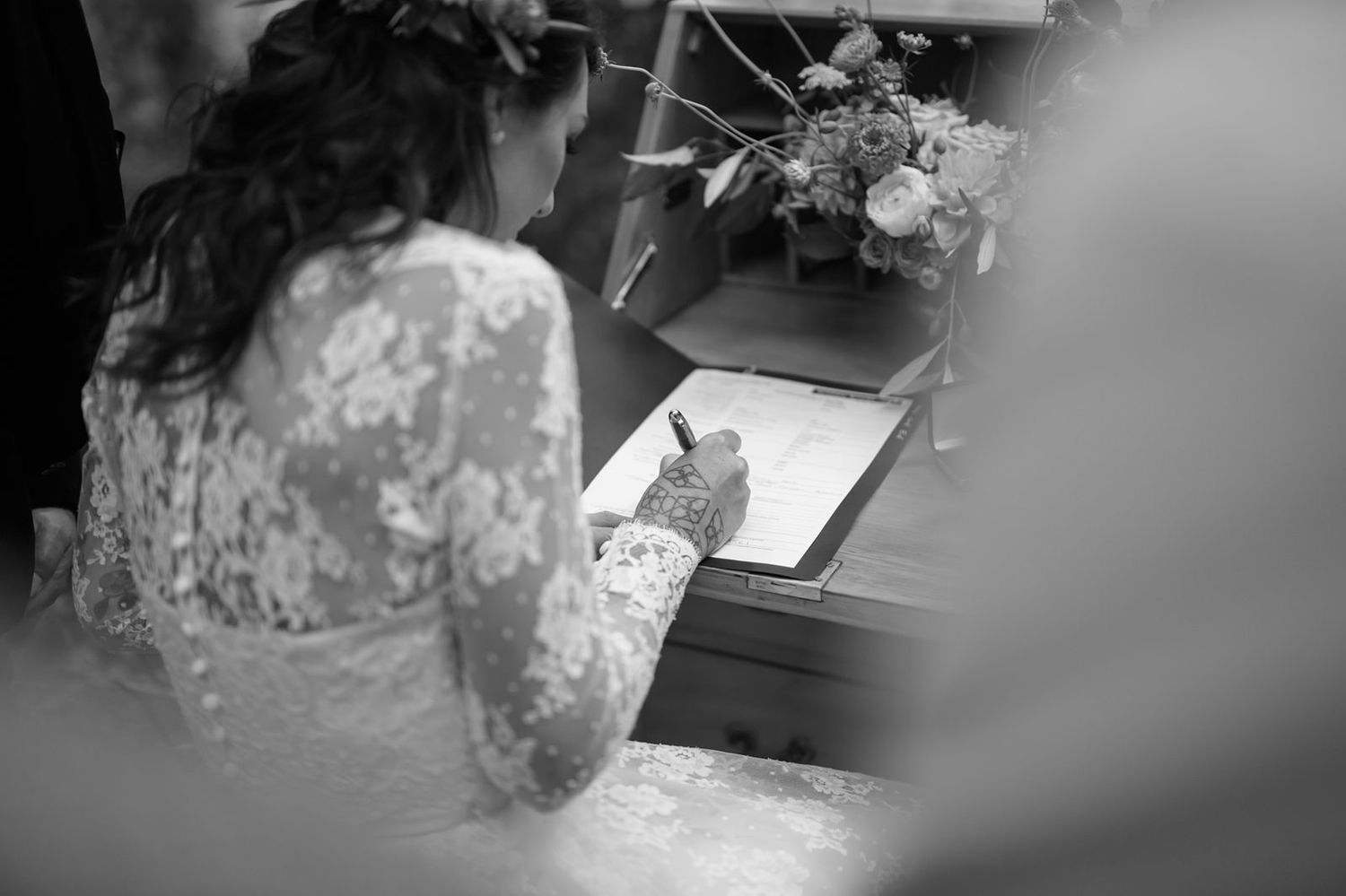 A person in lace writing on paper at a desk, photographed in black and white with soft focus.