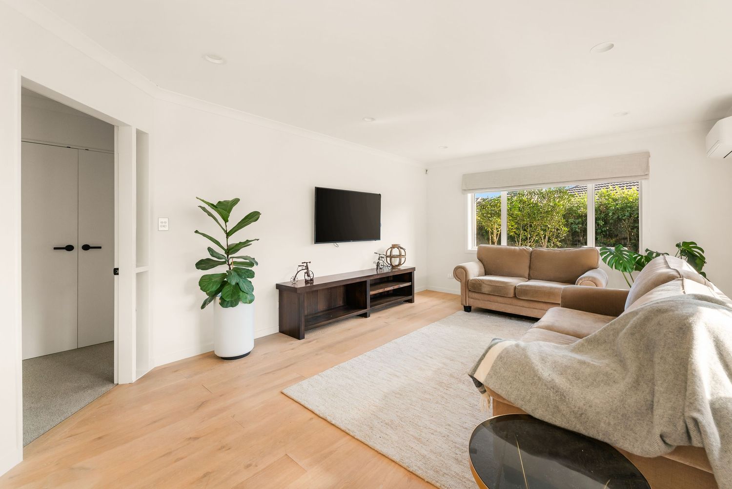 Modern living room with wood floors, mounted TV, black media console, tan sofa and potted plants by window.