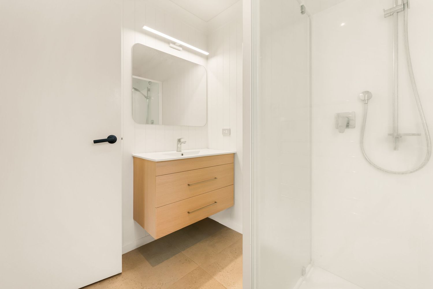 Clean modern bathroom with floating wooden vanity, white sink and glass shower enclosure.