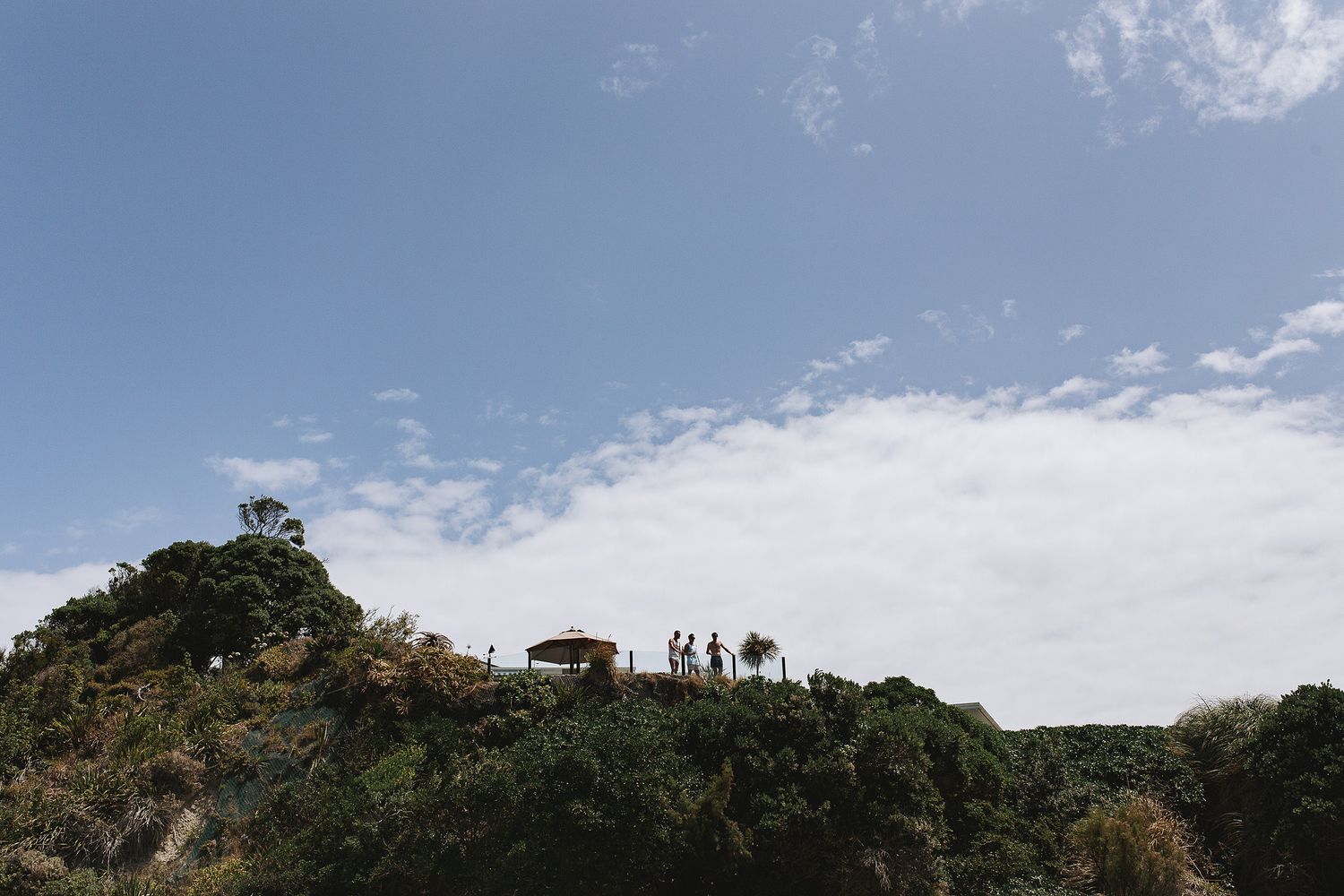 Silhouettes of people stand on a hilltop against a bright blue sky with scattered white clouds.