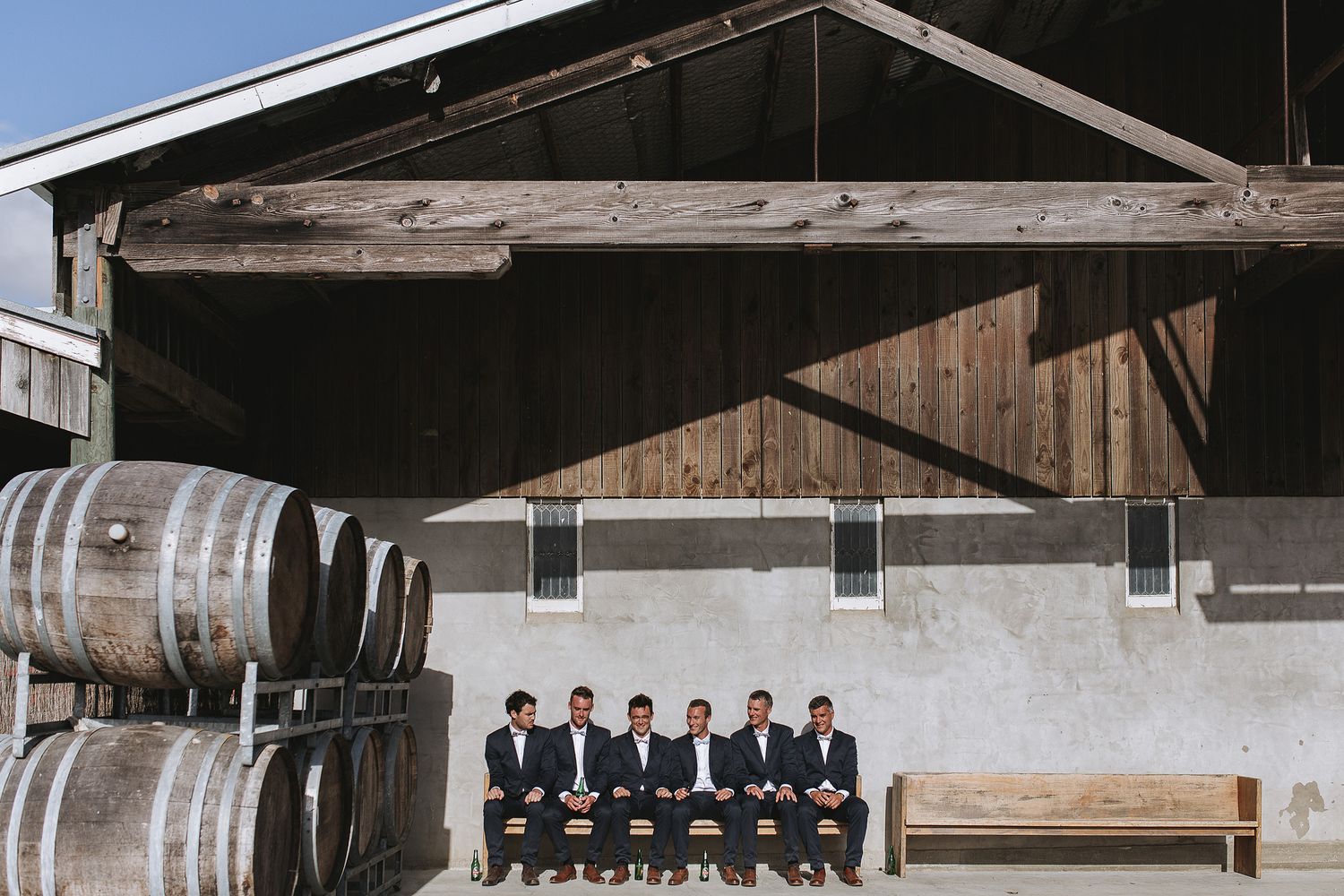 A group sits on a bench against a concrete wall with wooden wine barrels stacked nearby at a winery.