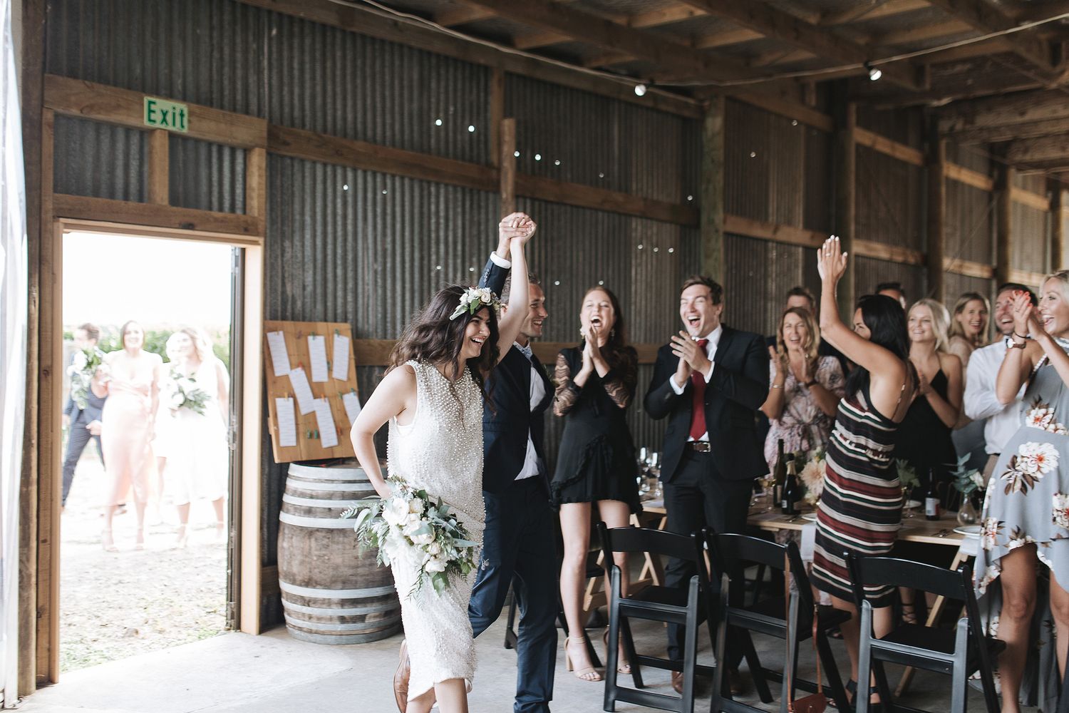A celebratory group of wedding attendees cheer and raise their arms in joy inside a rustic wooden barn venue.