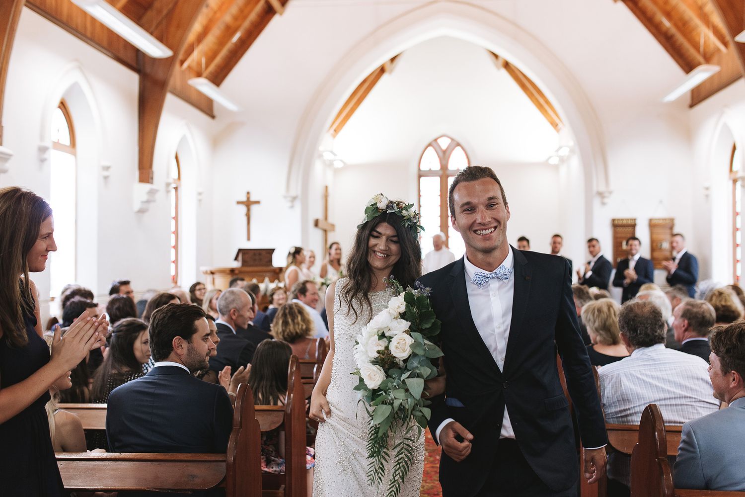 A bride and groom joyfully walk down the church aisle after their ceremony while guests look on from wooden pews.