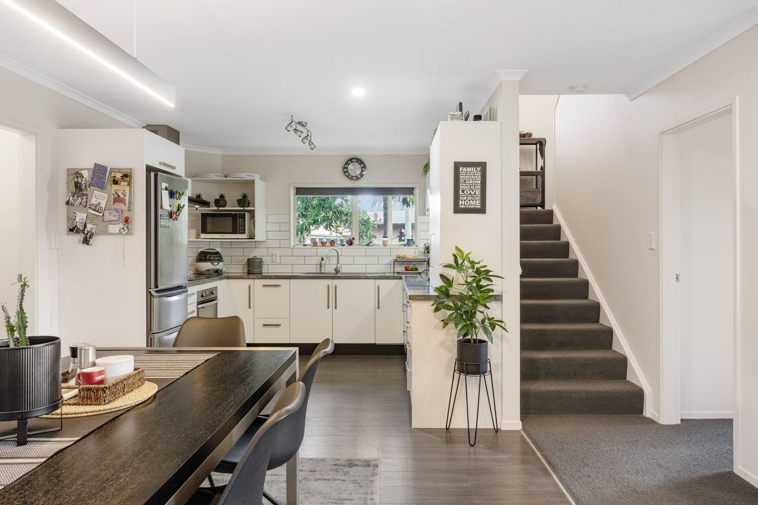 Modern white kitchen with dark flooring, wooden dining table, and staircase leading to upper level.