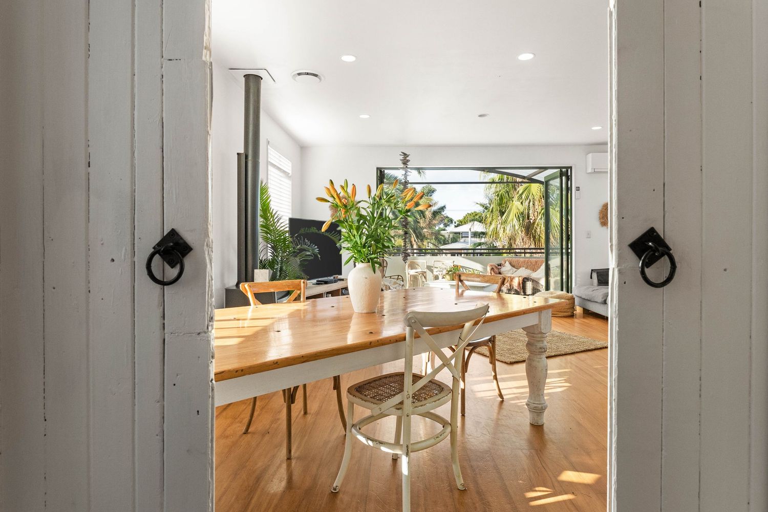 Bright modern dining room with wooden table, white chairs, and large windows overlooking tropical foliage.