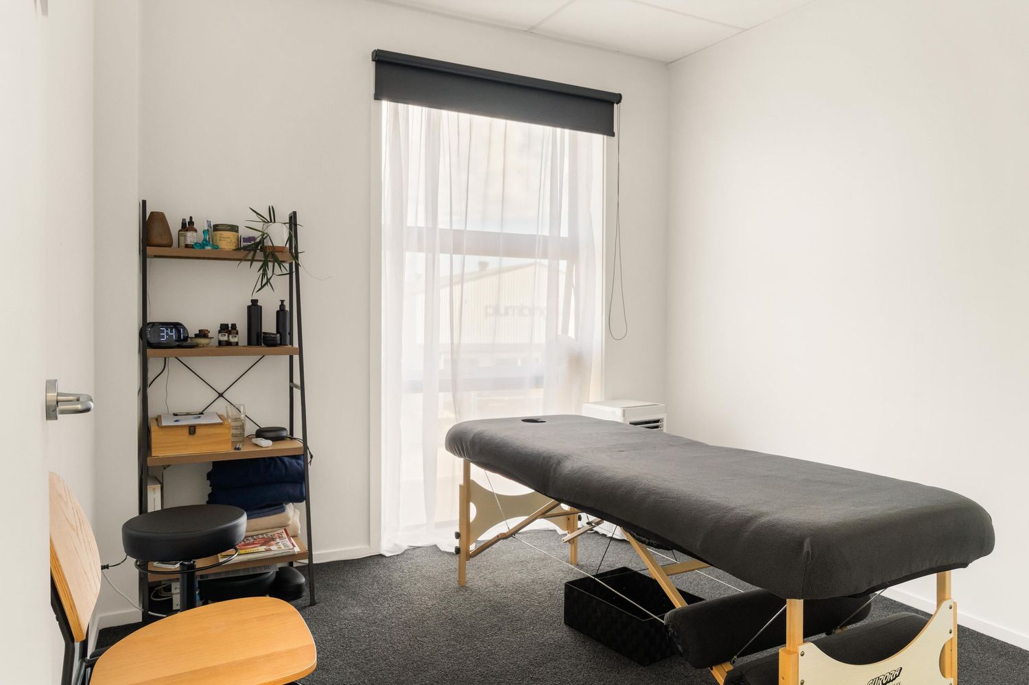 Minimalist white treatment room with massage table and wooden shelving units.