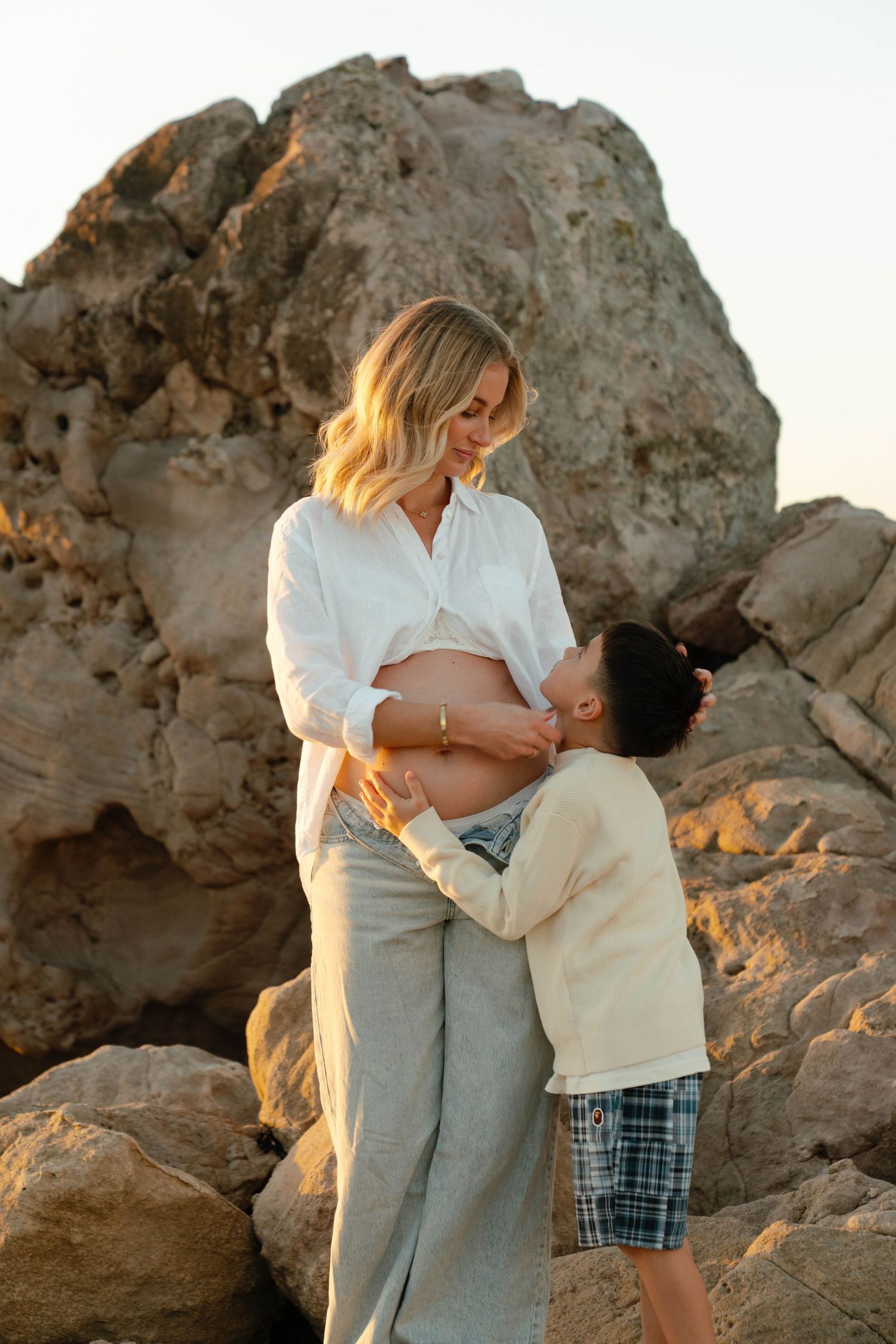 A child kisses a pregnant woman's belly outdoors near rocky cliffs at sunset. Photography by Bryony Alexander at Newfound.