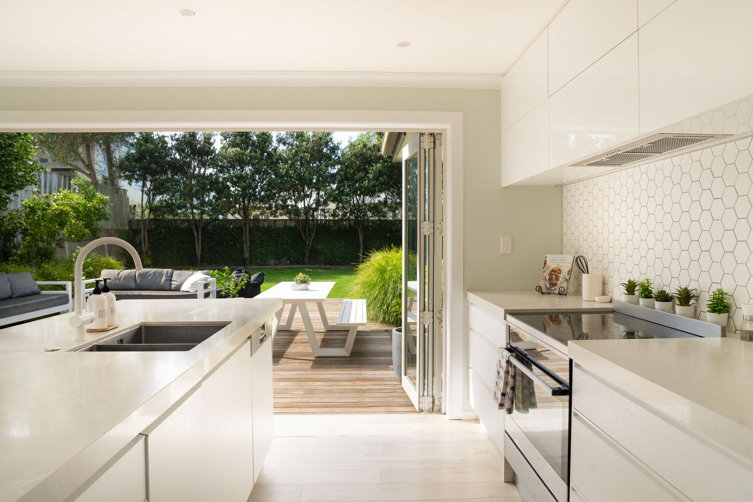 Modern white kitchen with bifold doors opening to a lush garden patio, featuring hexagonal tile backsplash and sleek cabinetry.