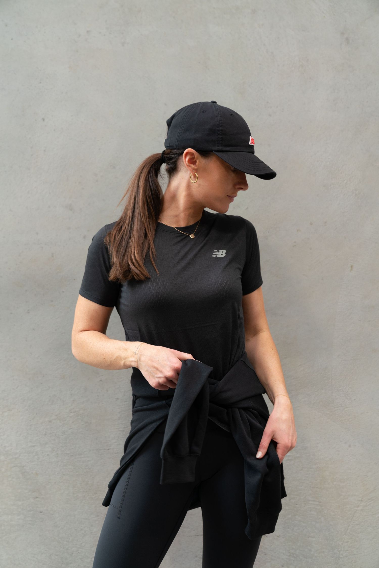 Woman in black New Balance t-shirt, cap, and leggings posing against a gray wall.