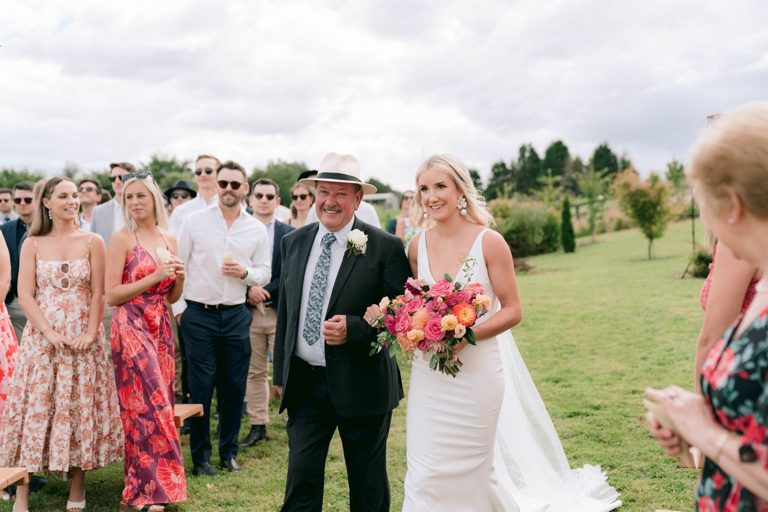 Wedding party celebrates on a lush green lawn during an outdoor summer ceremony with pink floral bouquet.