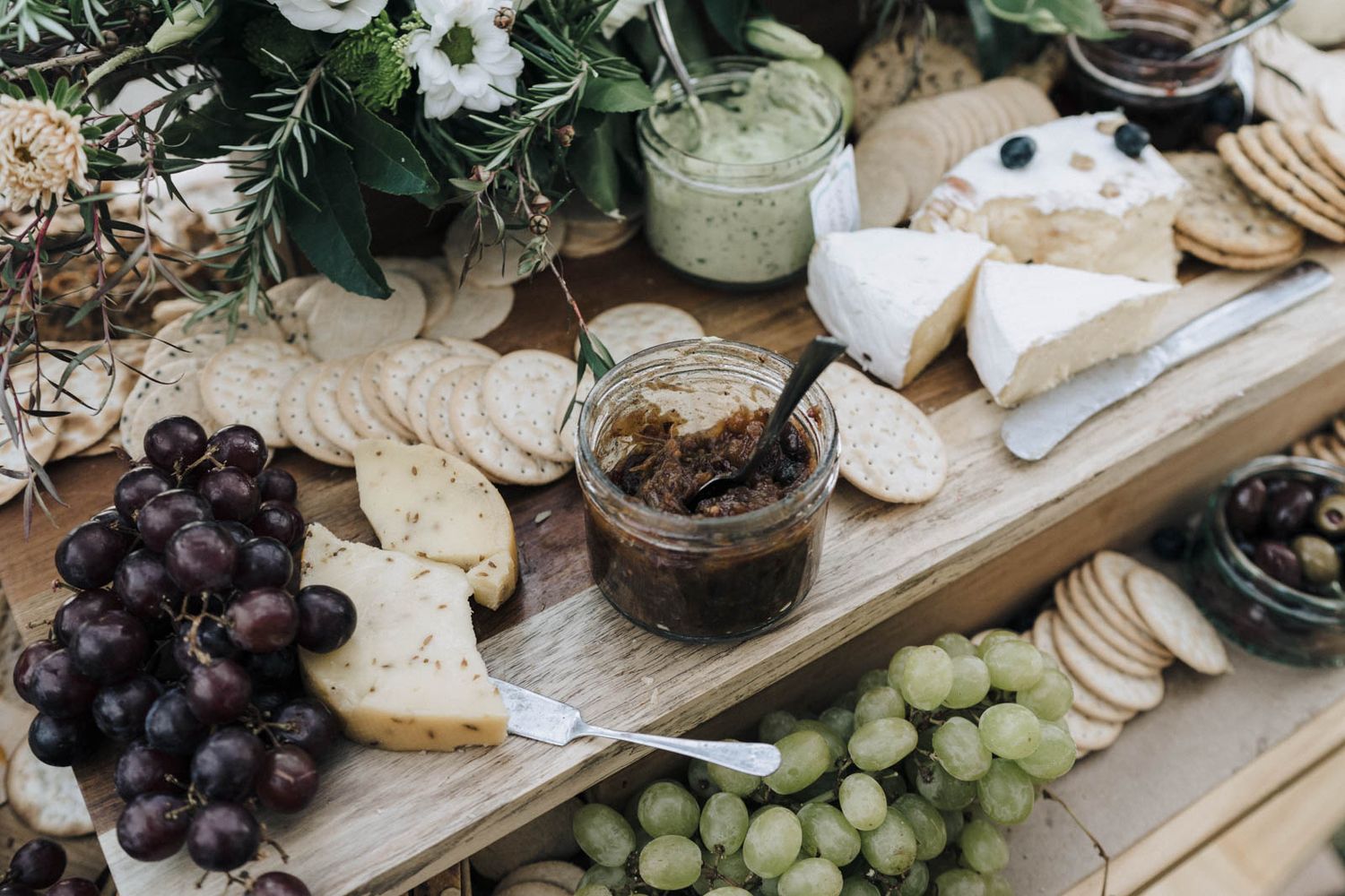 Rustic cheese and charcuterie board with grapes, crackers, jam, and flowers on a wooden serving plank.