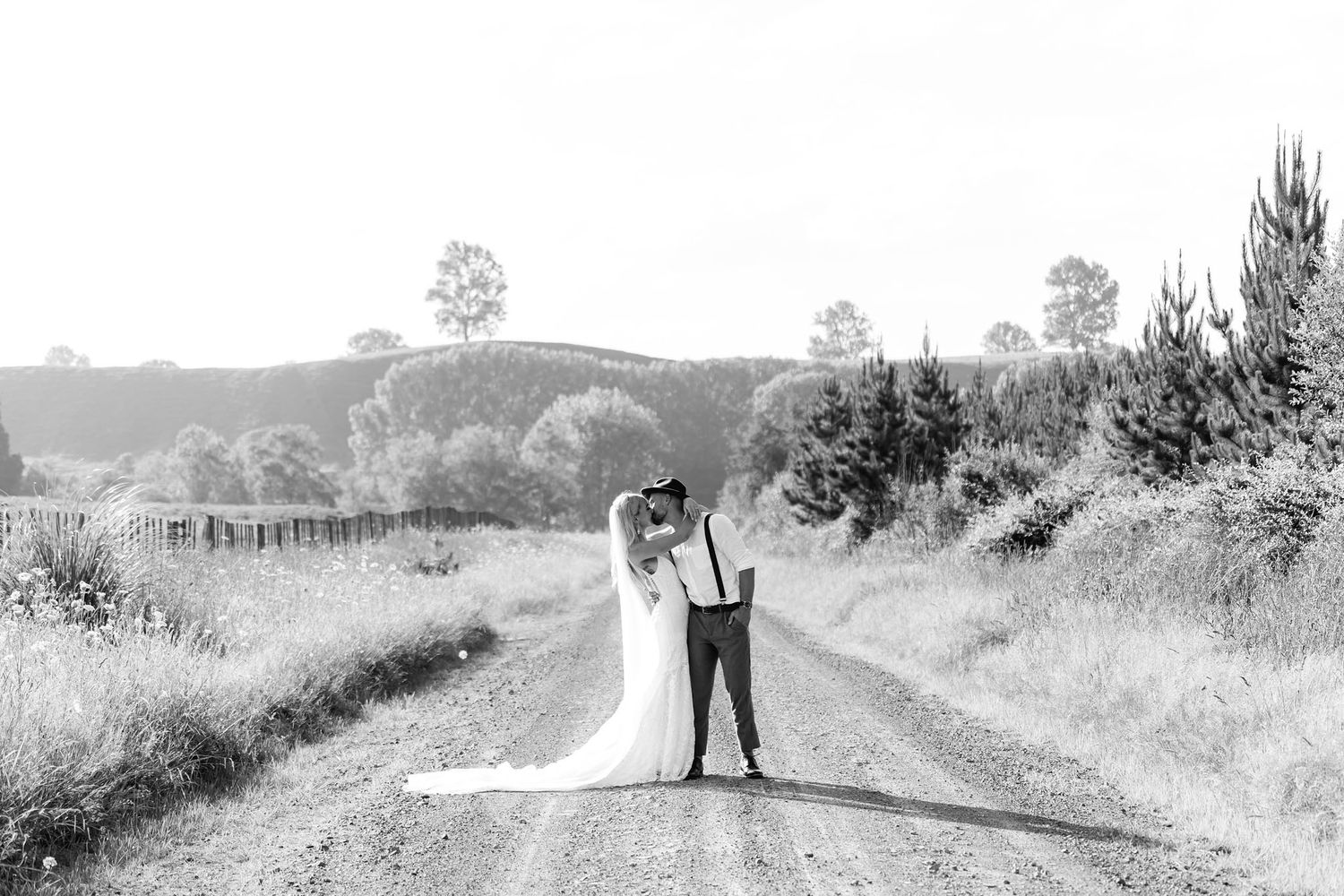 A couple in wedding attire stands together on a rural dirt road surrounded by rolling hills in black and white.