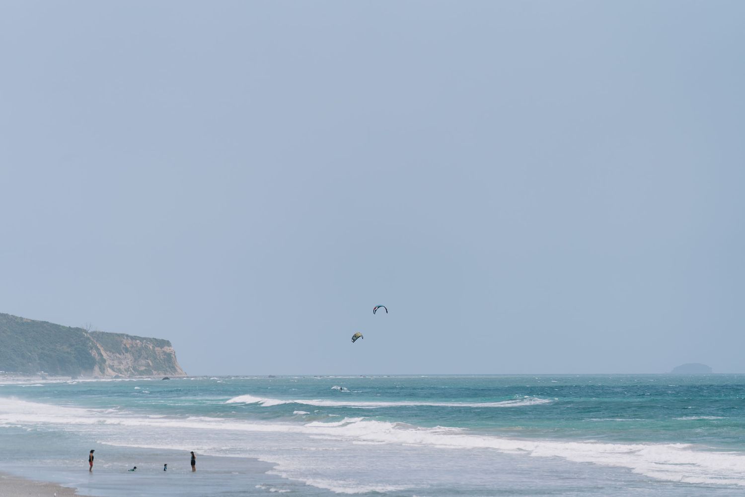 People walk on a misty beach with rocky cliffs and seabirds soaring over turquoise waves along the coastline.