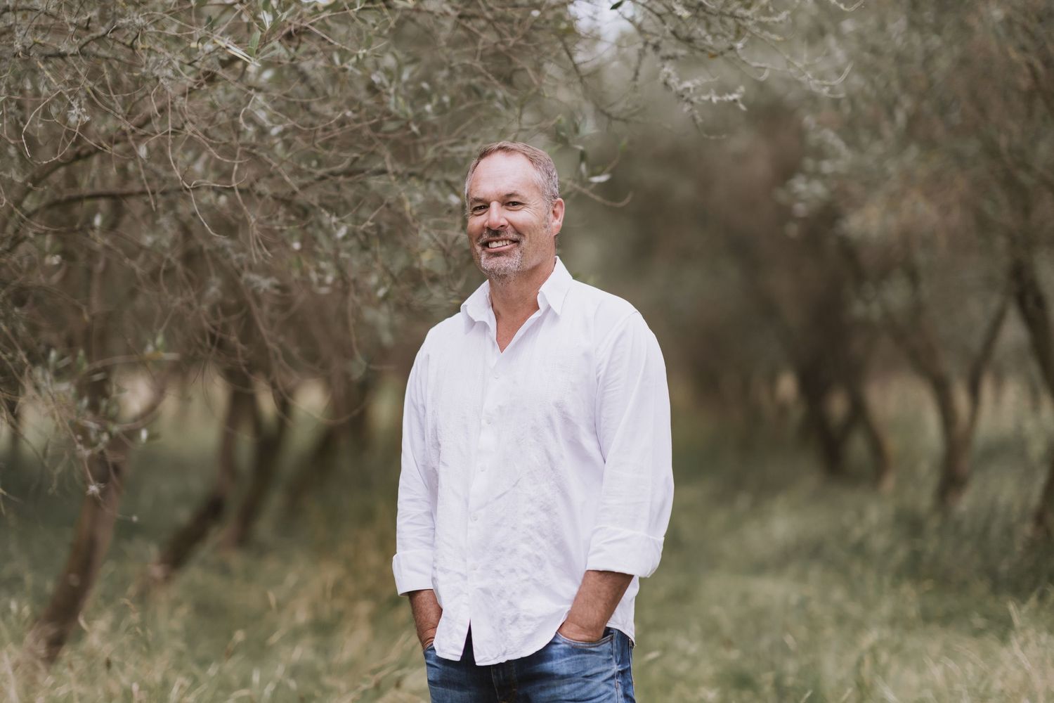 A relaxed outdoor photo in an olive grove showing someone in a white shirt and jeans with a gentle expression.