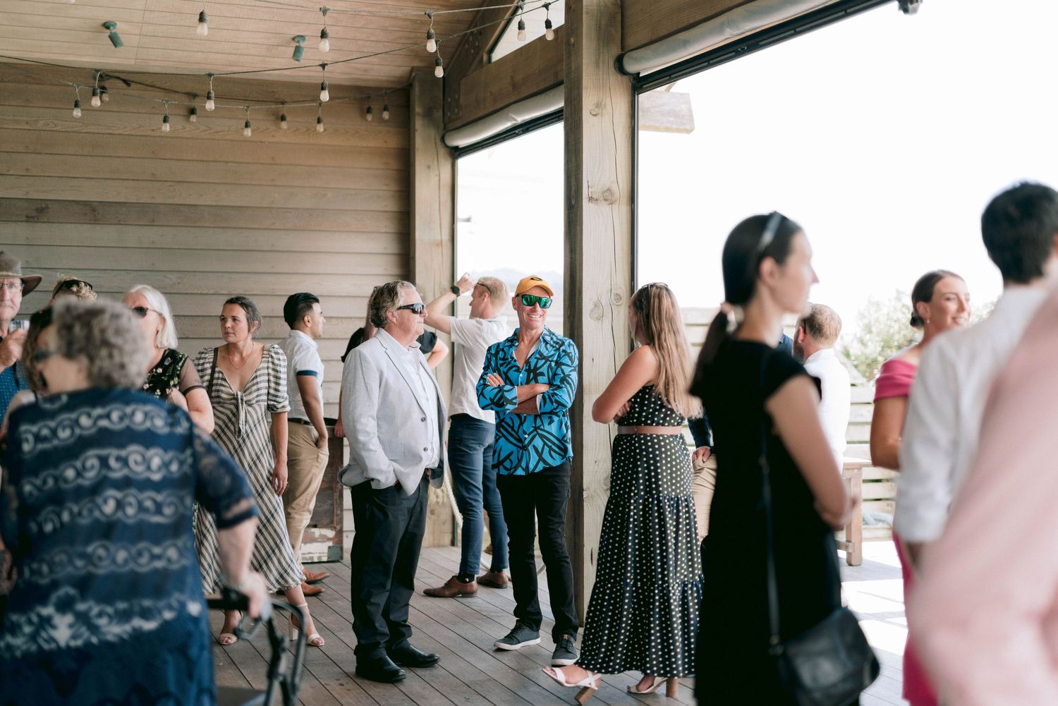 Wedding guests gather on a covered outdoor deck during a casual reception celebration.