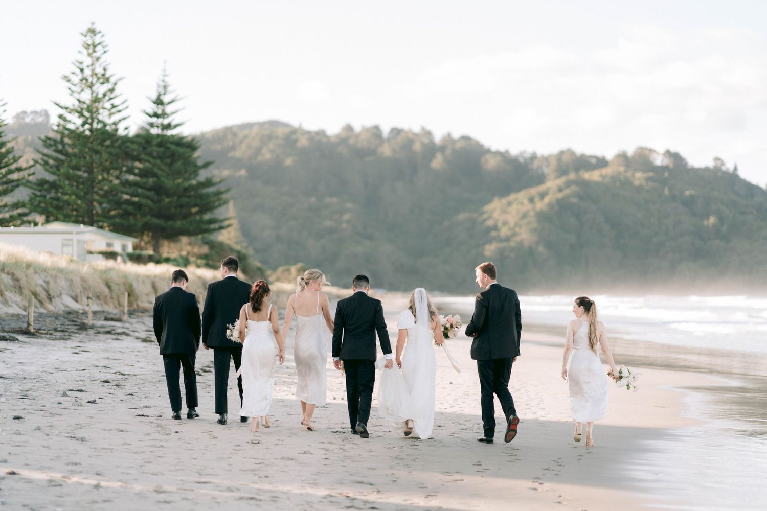Wedding party walking along a scenic beach with mountains and pine trees in background at sunset.
