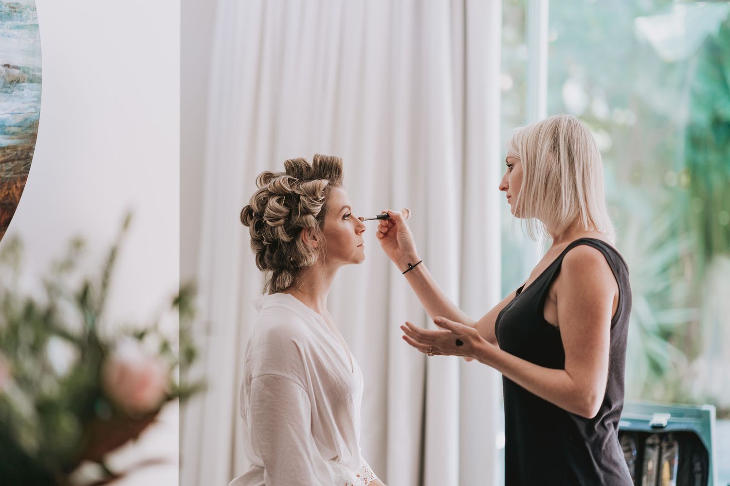Professional makeup artist applies cosmetics to bride during wedding day preparation in bright white room.
