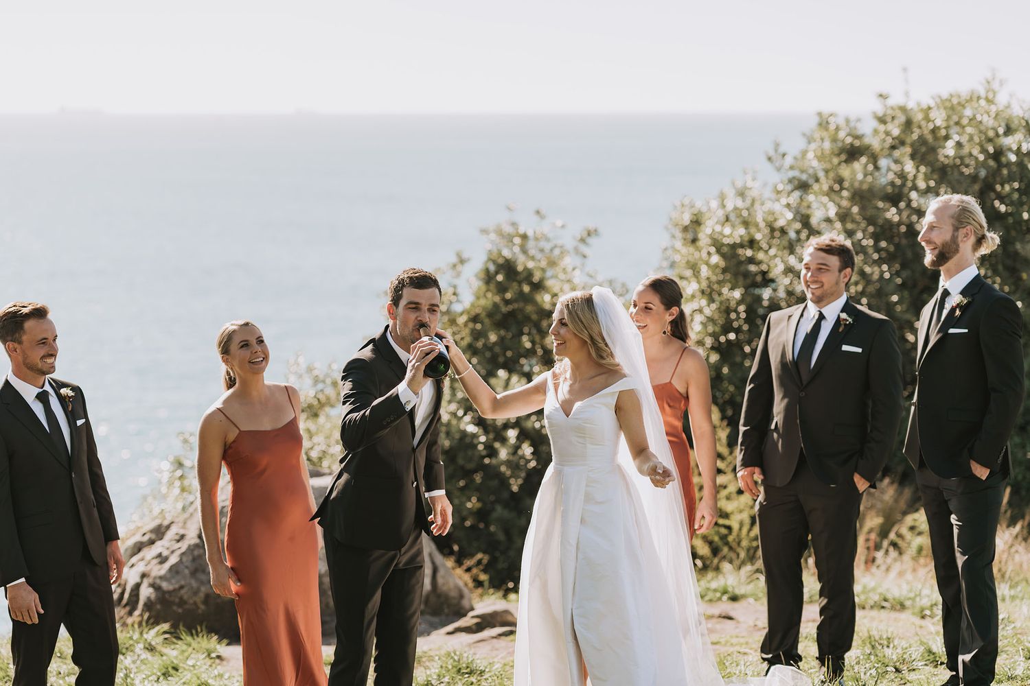 A wedding party stands together in a line on a hilltop during a sunny outdoor ceremony with scenic mountain views.