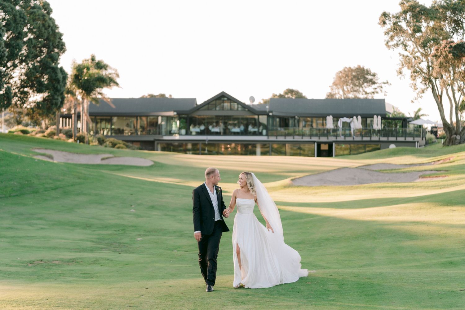 Bride and groom walking hand in hand across a lush green golf course during sunset.