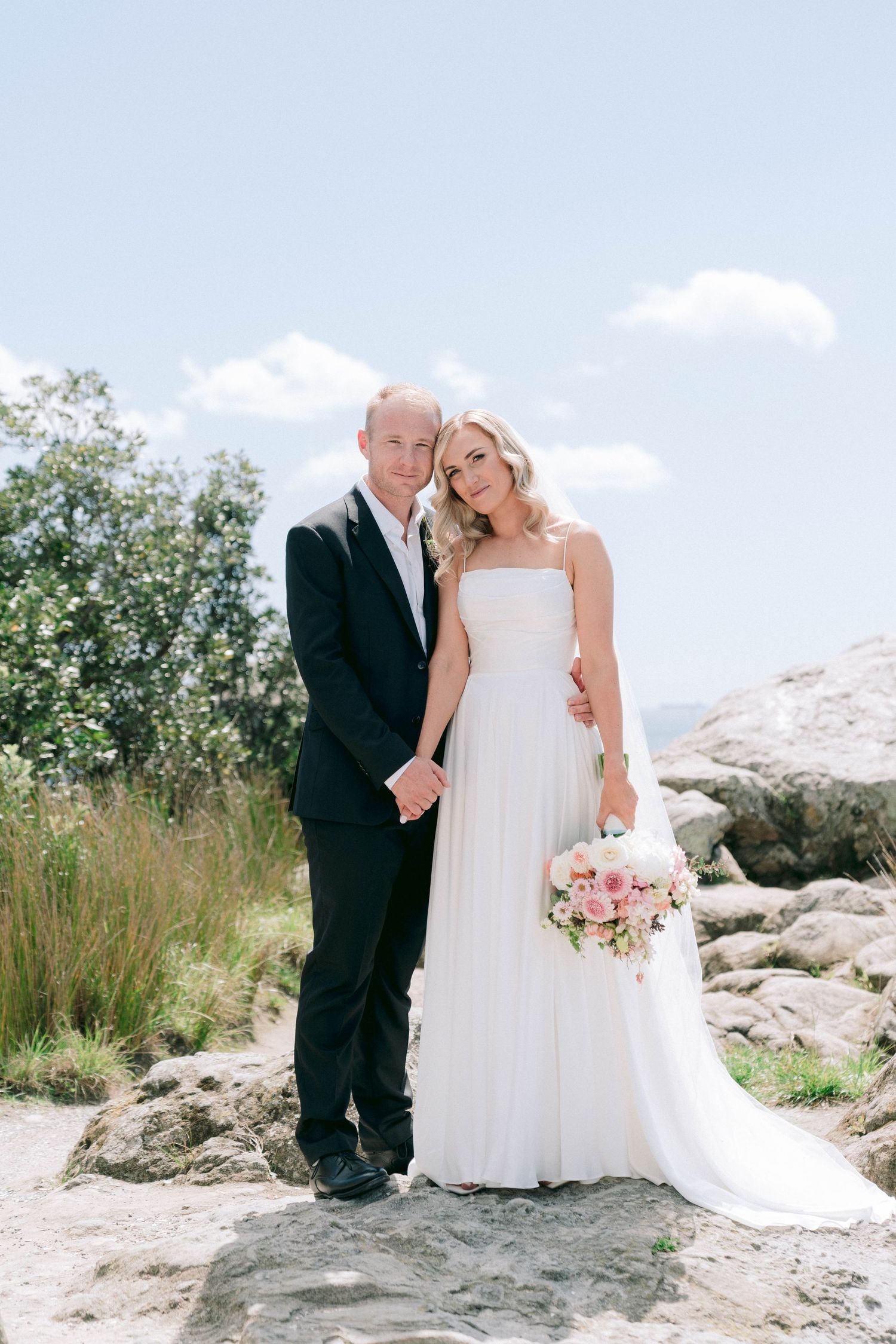 A wedding couple poses together on rocky terrain with mountains and blue sky in the background.