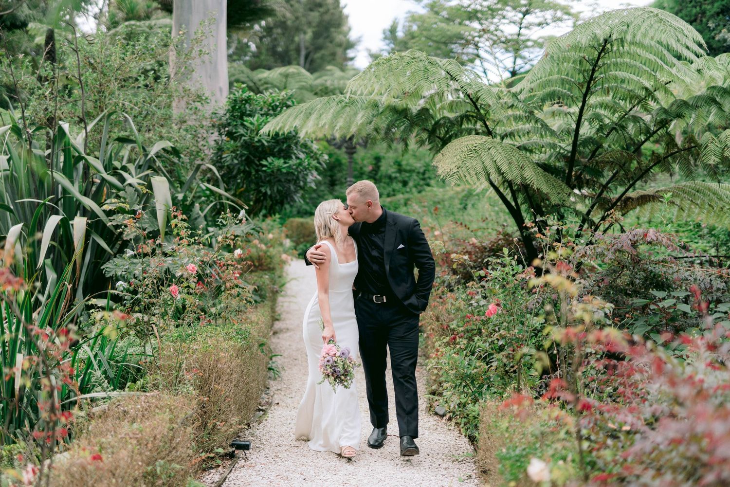 A boardwalk path winds through a garden setting with blooming flowers and ferns creating a romantic atmosphere.