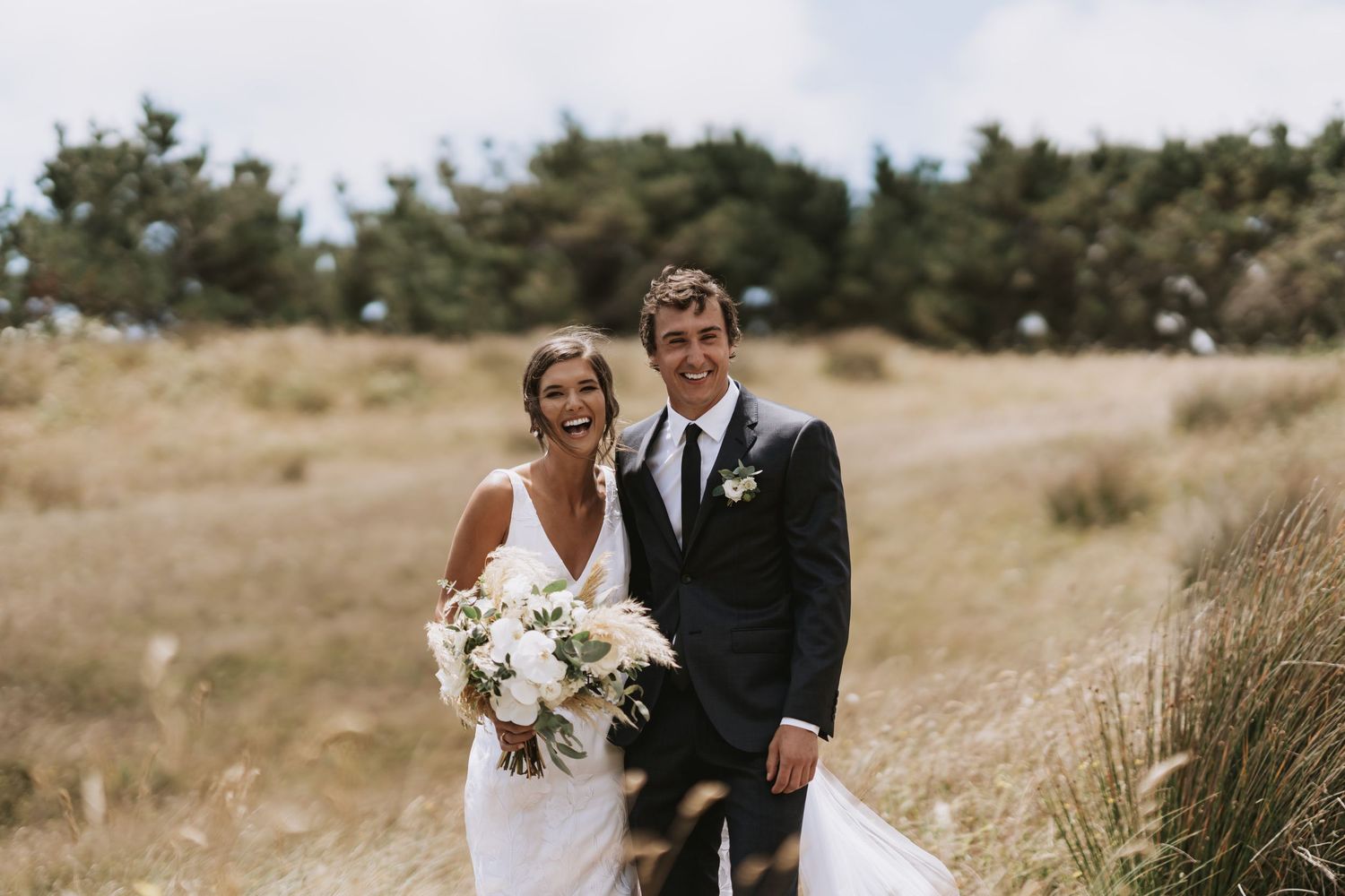 A newlywed couple stands together in a grassy coastal field holding a white floral bouquet on their wedding day.