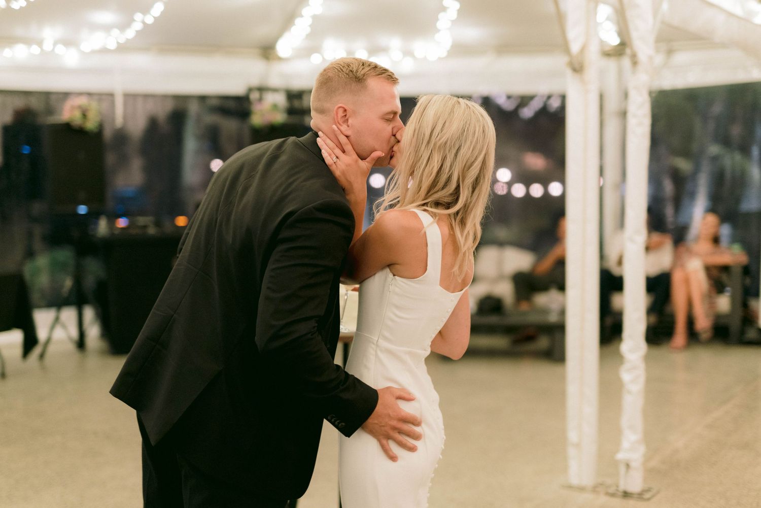 A romantic kiss between a couple at their evening wedding reception under a white tent with twinkling lights.