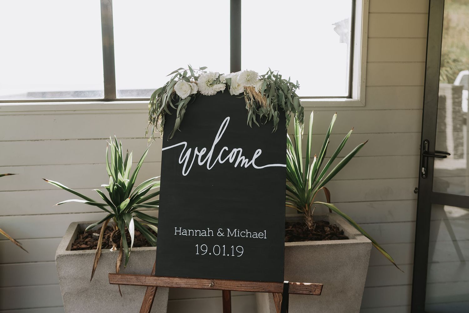 A dark welcome sign for Hannah and Michael's wedding with white lettering and floral decorations, flanked by potted plants.