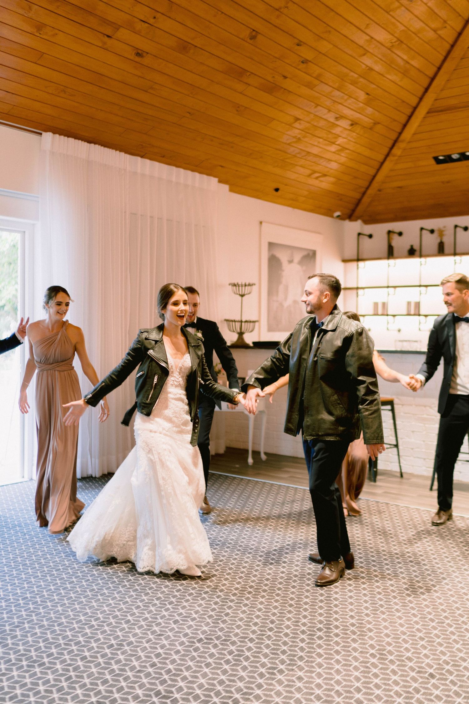 Wedding party dancing in a reception venue with wooden ceiling and leather jackets.