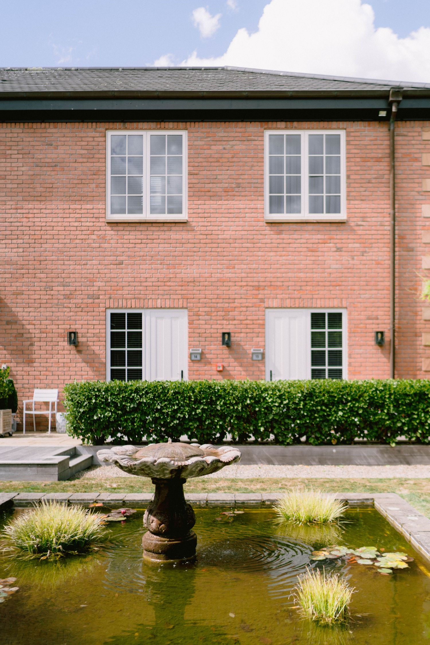 A tranquil garden pond with ornamental grasses and water features against a brick building exterior.