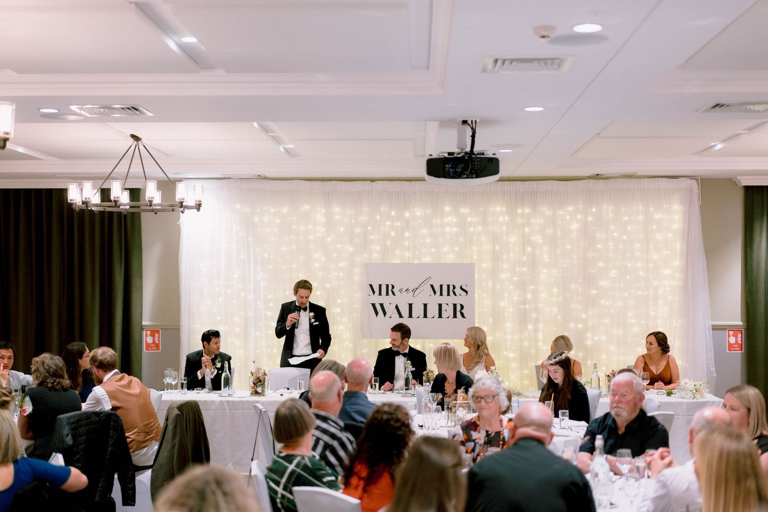 Large indoor event with banquet tables and a panel presentation setup featuring a light colored backdrop in a conference room.