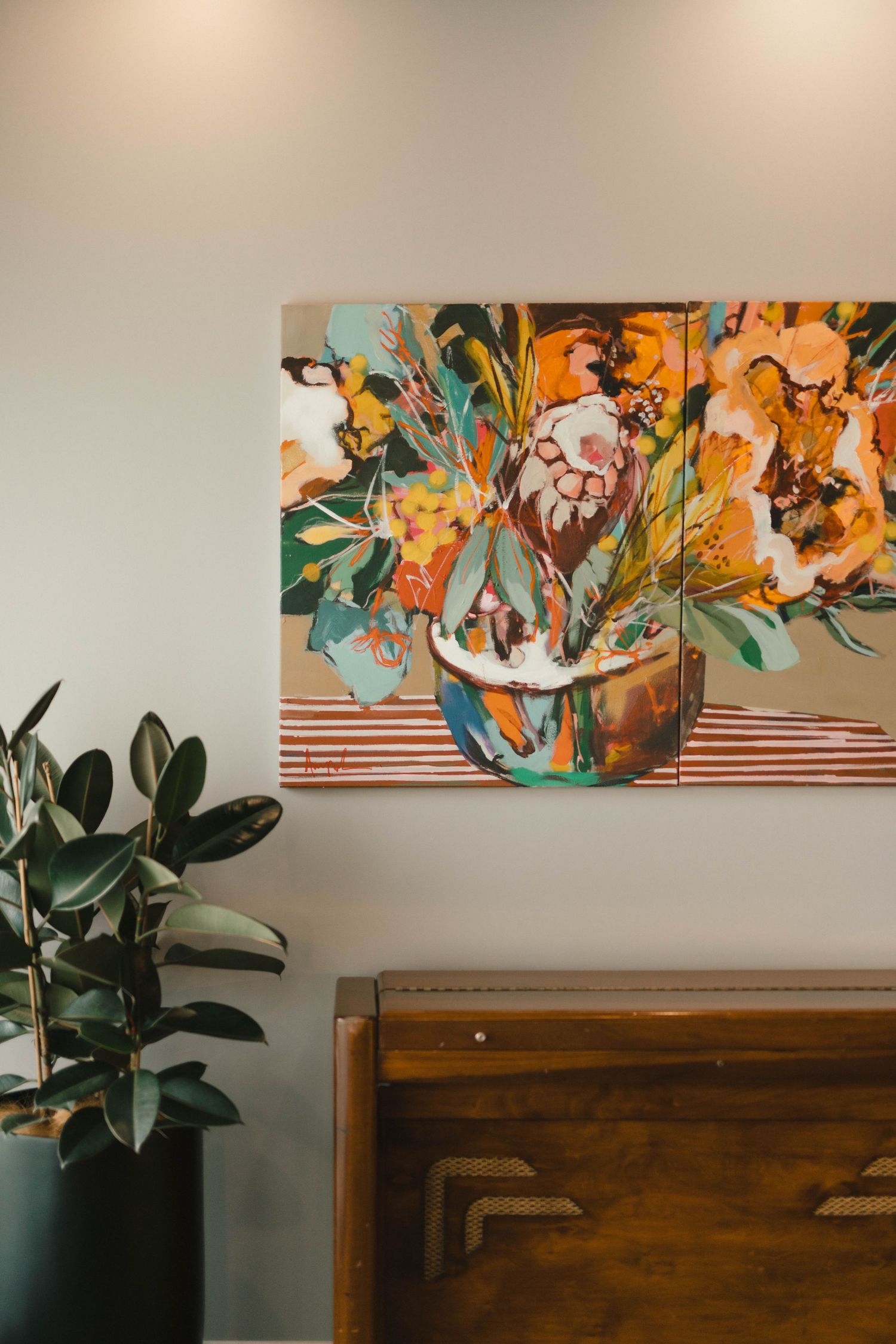 Colorful abstract floral painting hanging above wooden furniture with a potted plant nearby.