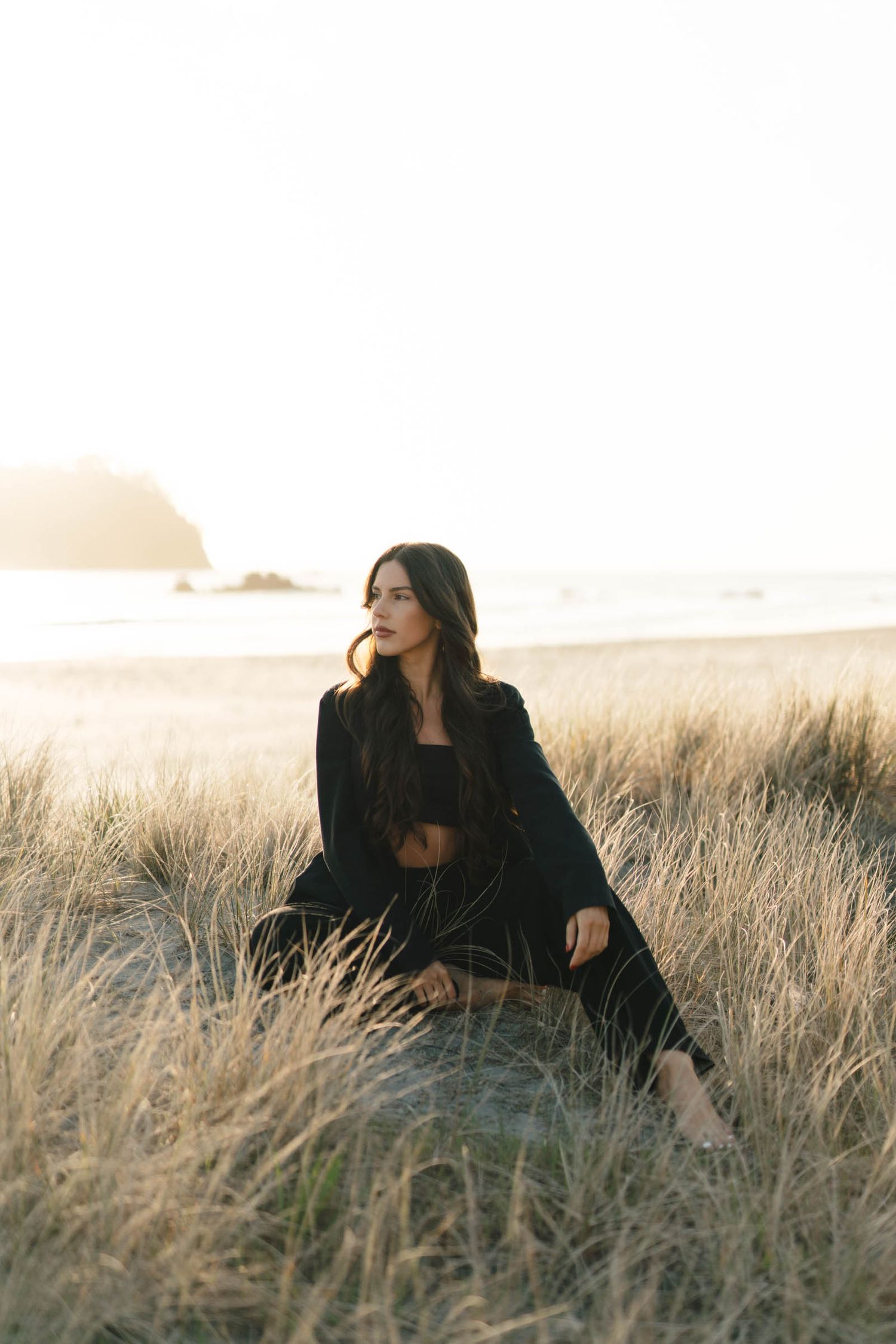 Figure in dark outfit sits thoughtfully among coastal grass at golden hour.