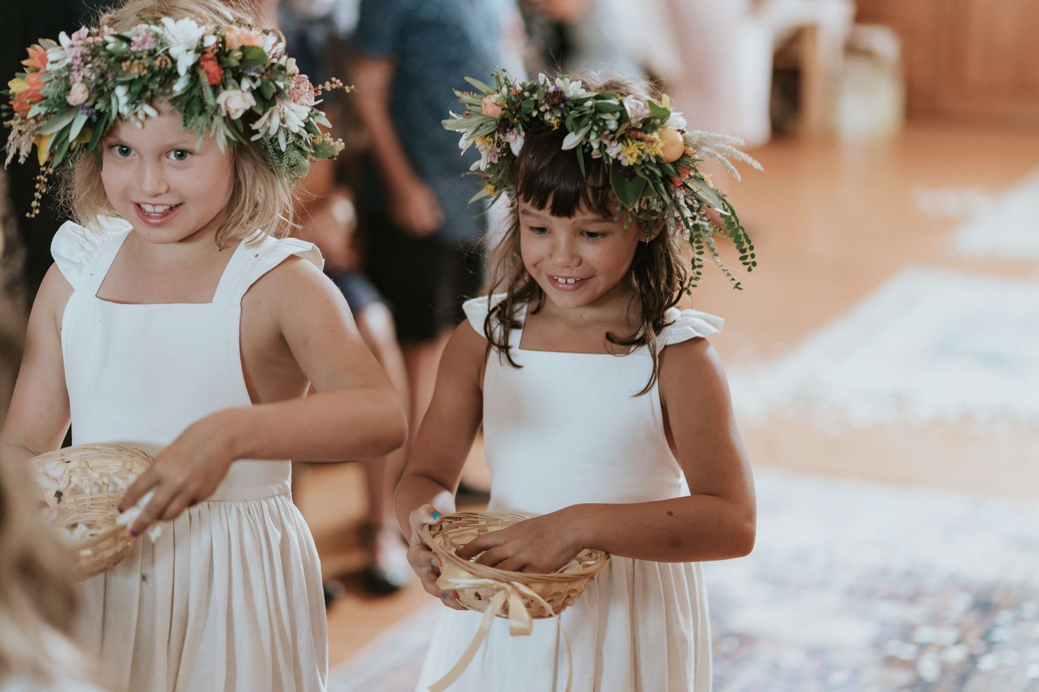 Two flower girls in white dresses and floral crowns smile while holding petals at a wedding ceremony.