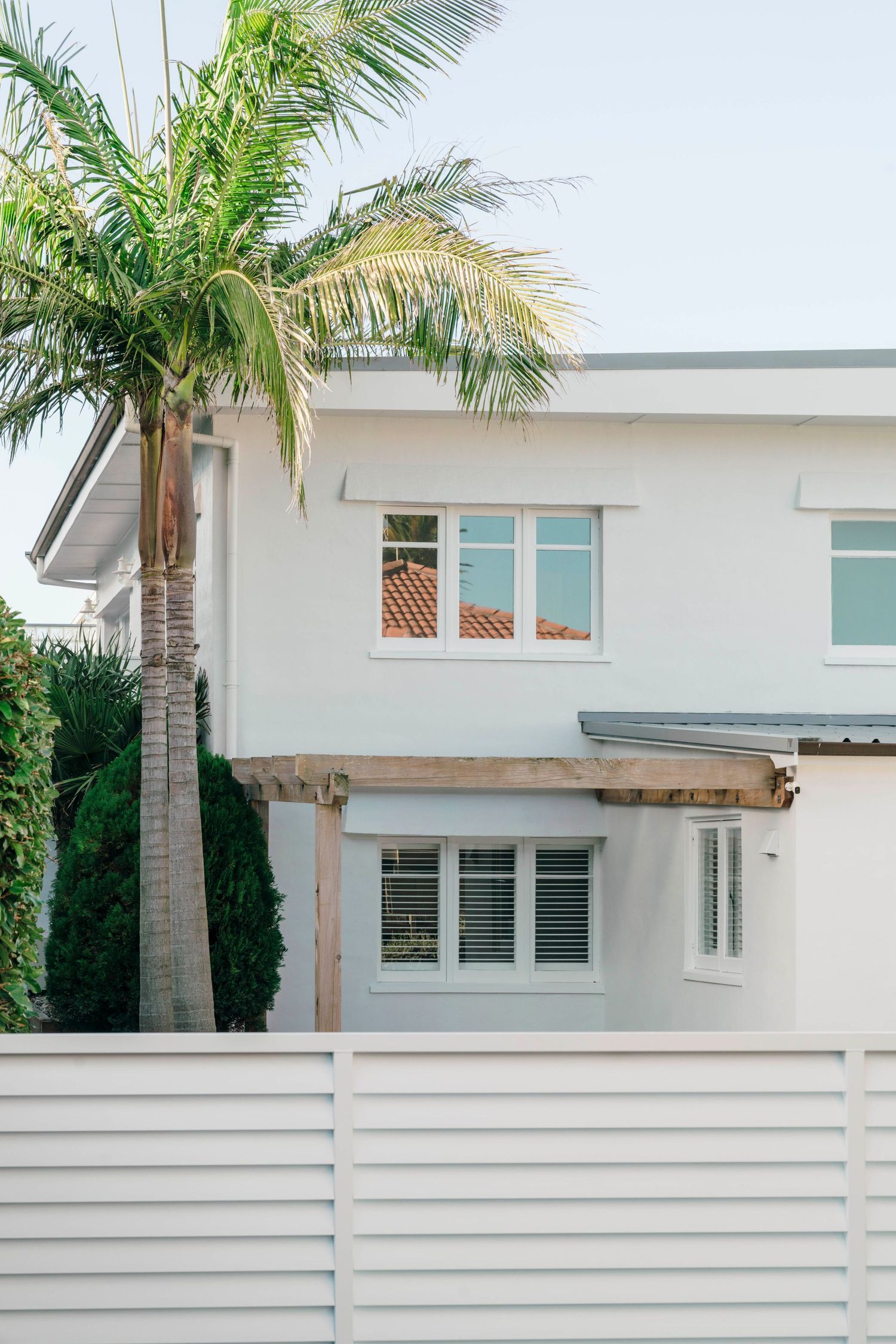 Modern white two-story house with palm tree and horizontal fence in tropical setting.