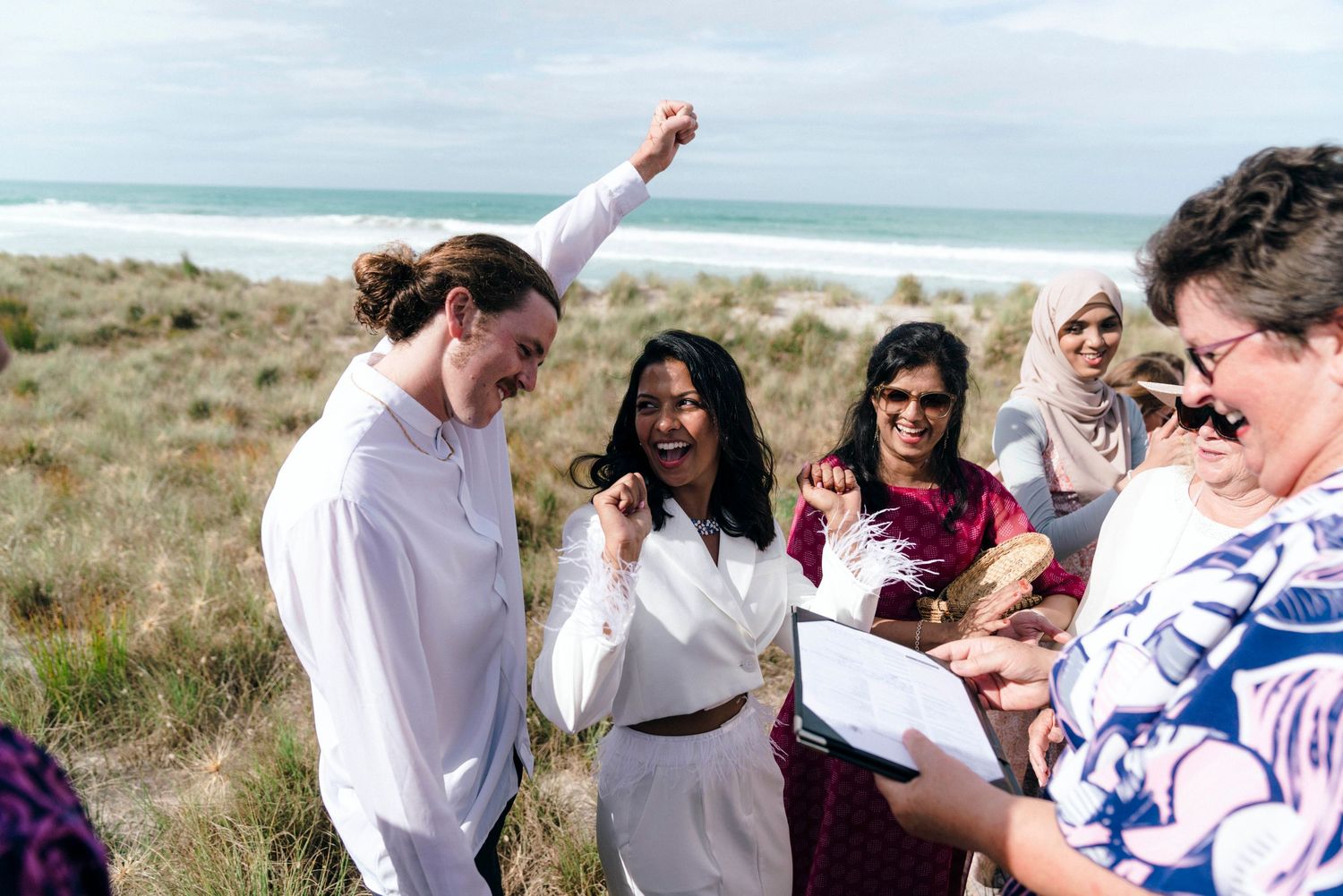 Group celebrates with raised arms during a seaside gathering in grassy dunes.