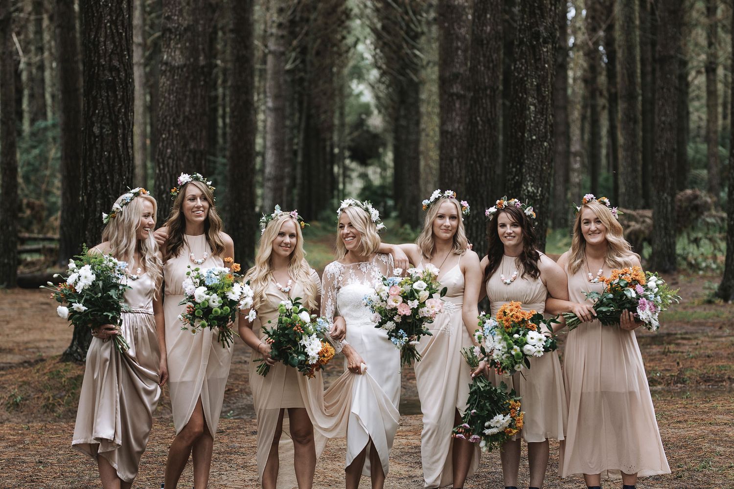 A group of women in neutral dresses and floral crowns stand together holding bouquets in a pine forest setting.