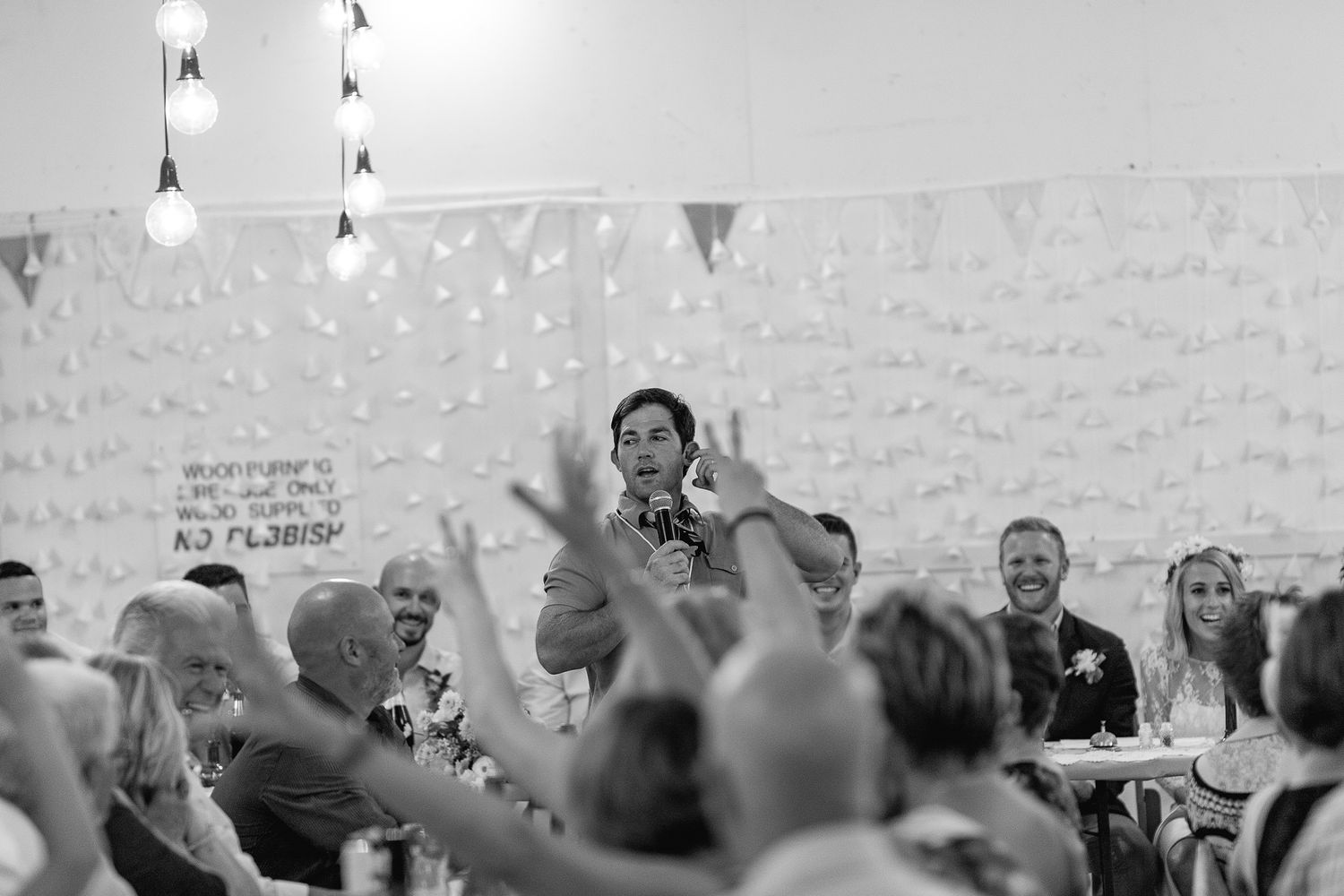 A lively wedding reception crowd raises their hands in celebration as speakers stand at the head table in black and white.