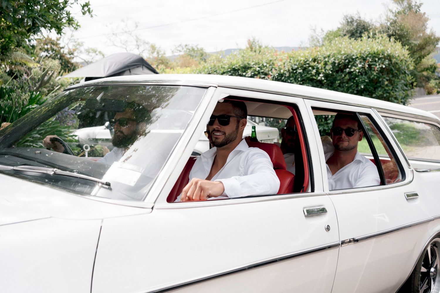 Vintage white car on sunny day with occupants wearing sunglasses traveling through green scenery.